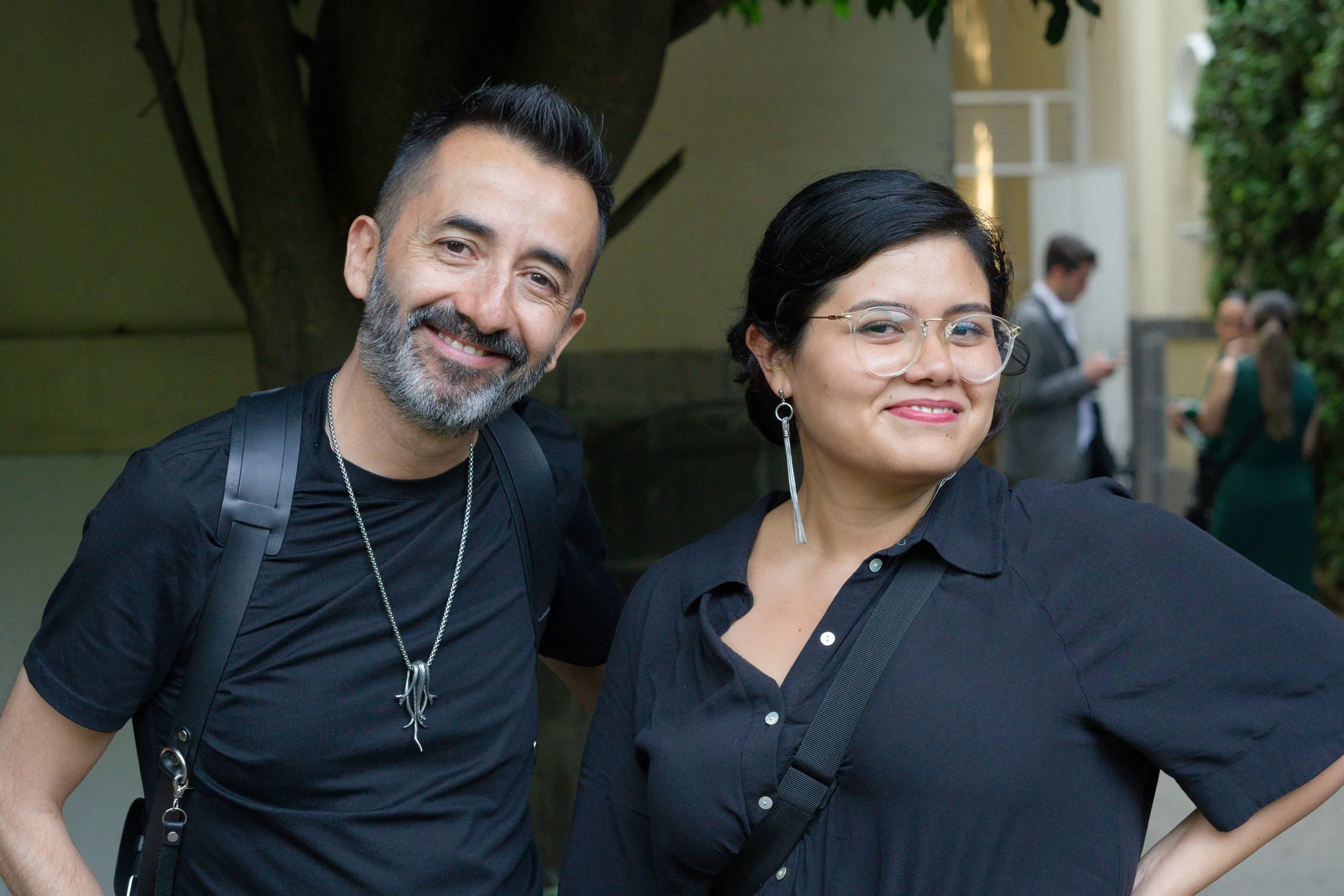 A smiling middle-aged man with a beard and a woman wearing glasses, both photographers dressed in black, standing outdoors with other people in the background.