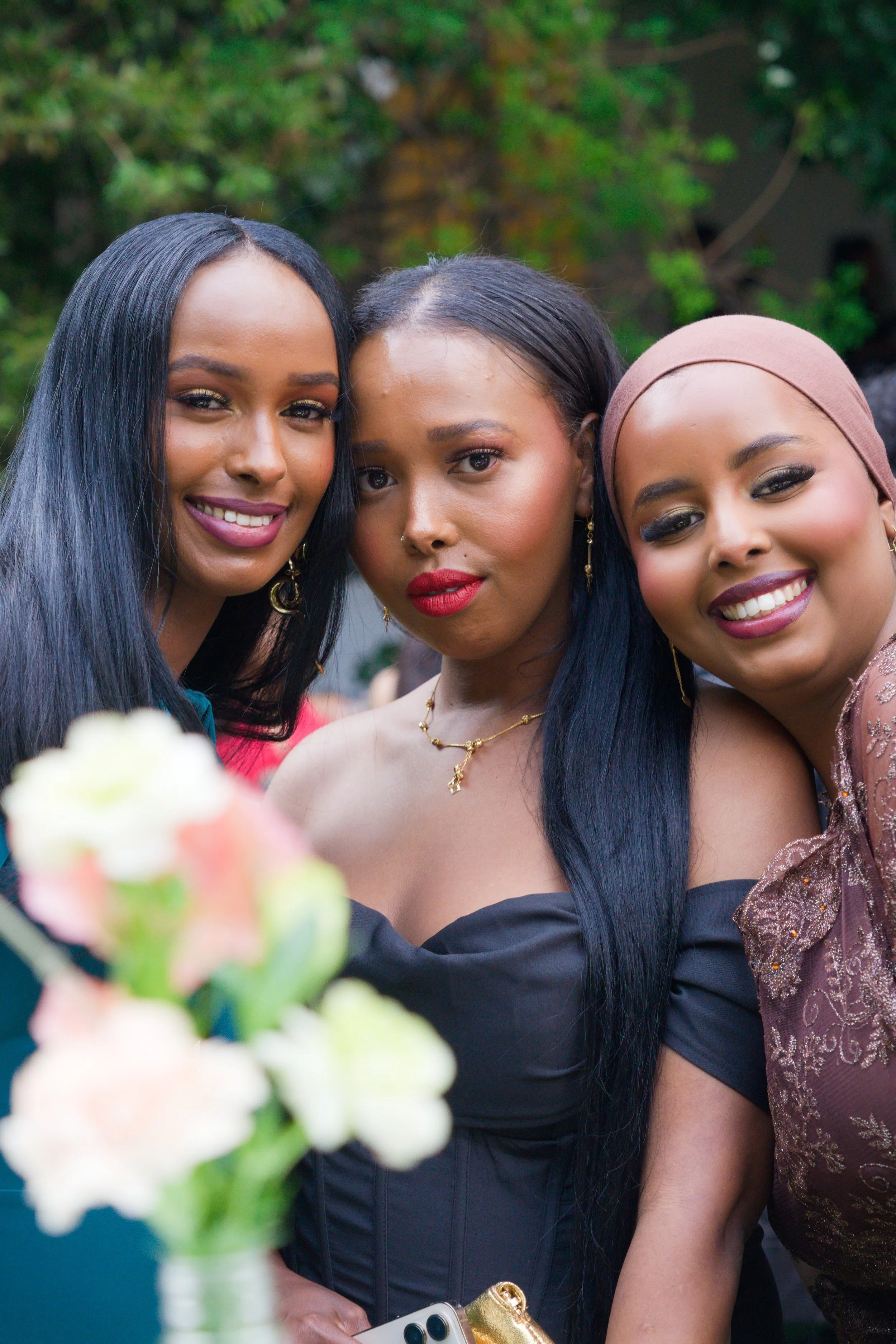 Three women in elegant attire smiling outdoors with a blurred flower bouquet in the foreground.