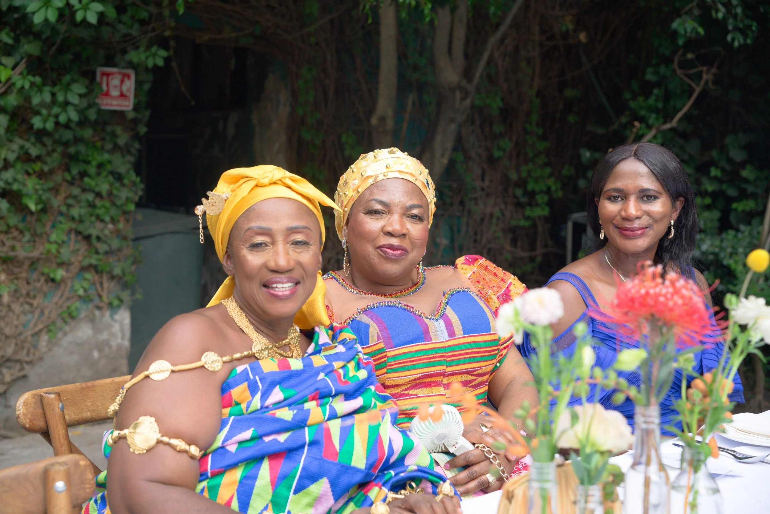 Three women wearing colorful traditional African clothing and headwraps sitting at a table with flowers.