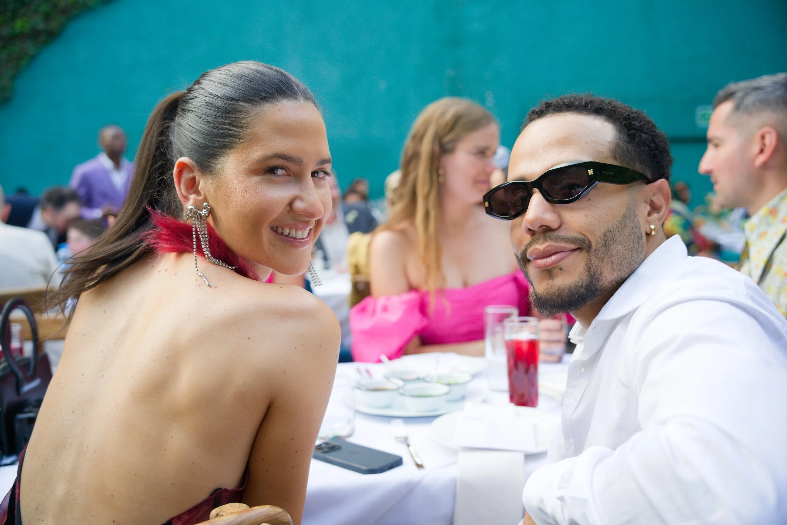 Two people smiling at a gathering, with tables set with drinks and dishes, other guests in the background, outdoors with a teal wall behind.