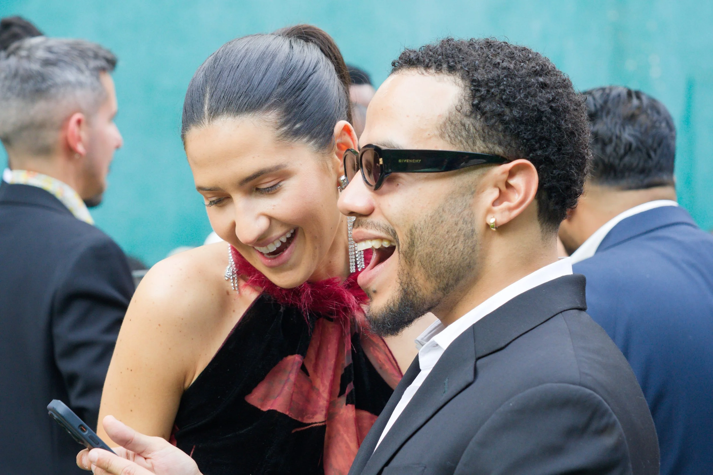 Two people, a woman and a man, smiling and looking at a smartphone, at a formal event with others in the background.