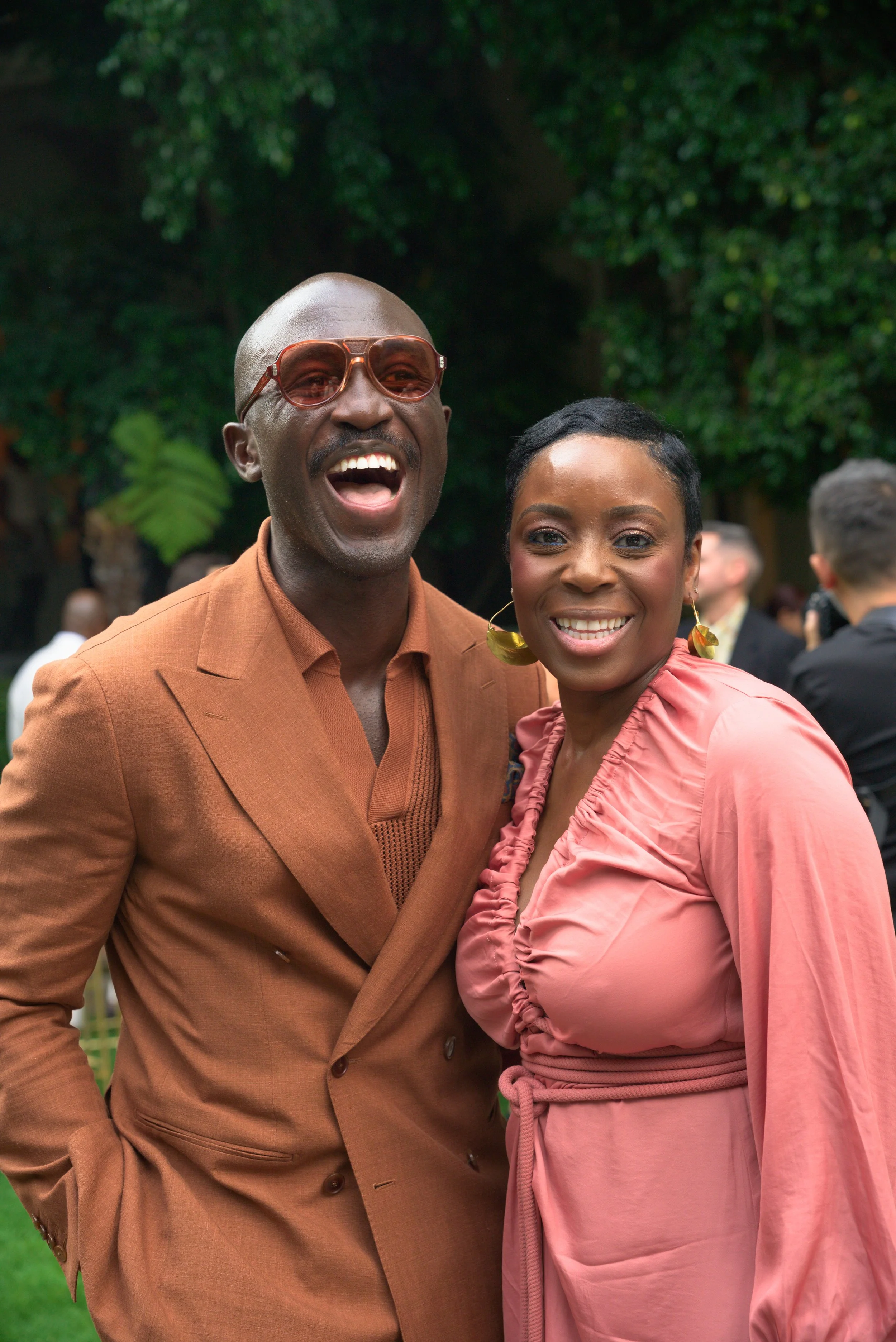 A smiling man wearing a brown suit and sunglasses, standing next to a smiling woman in a pink dress with earrings, at an outdoor event with greenery and other people in the background.