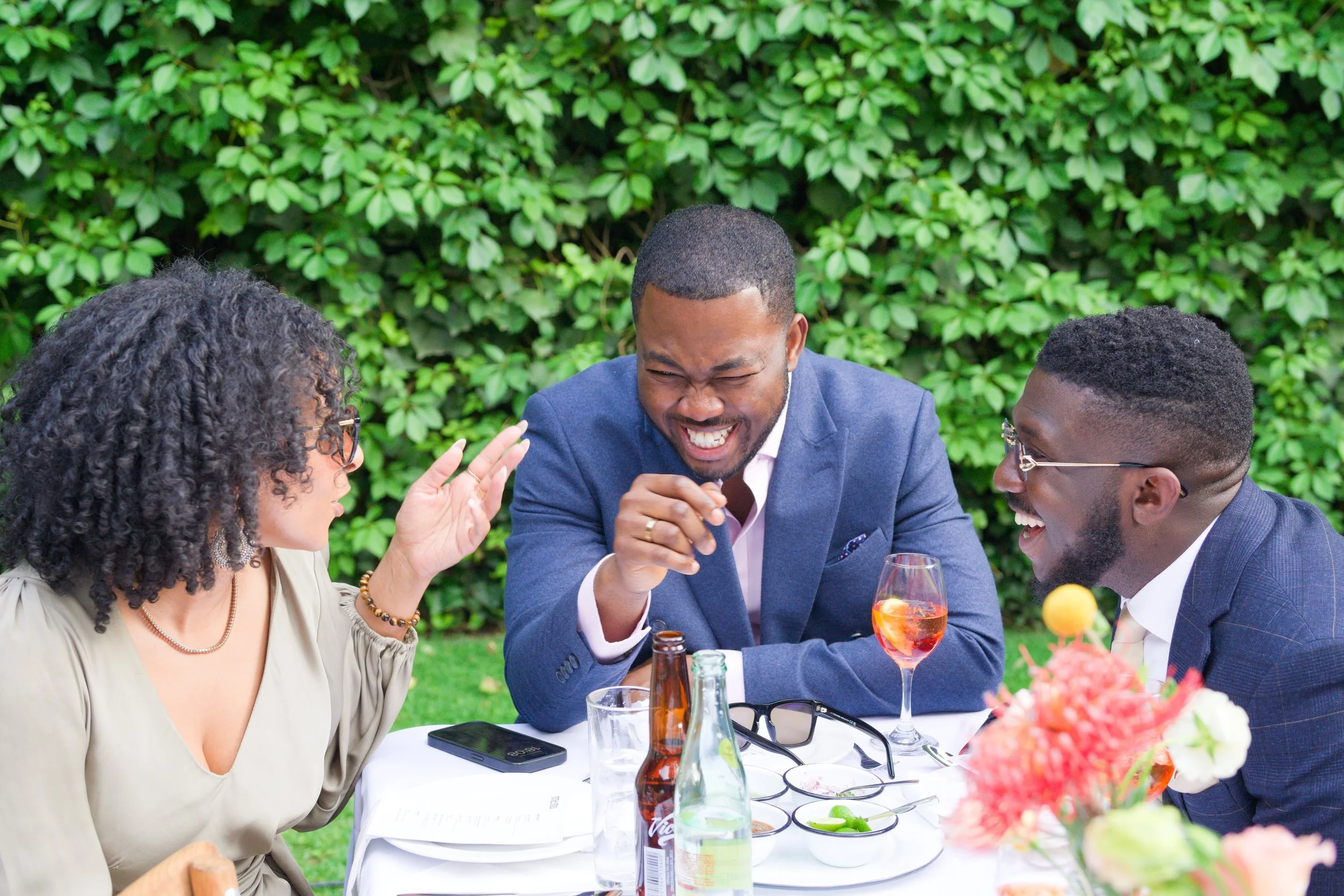 Three people sitting at a table outdoors, engaging in a lively conversation, smiling and laughing, with drinks and food on the table and a background of green foliage.