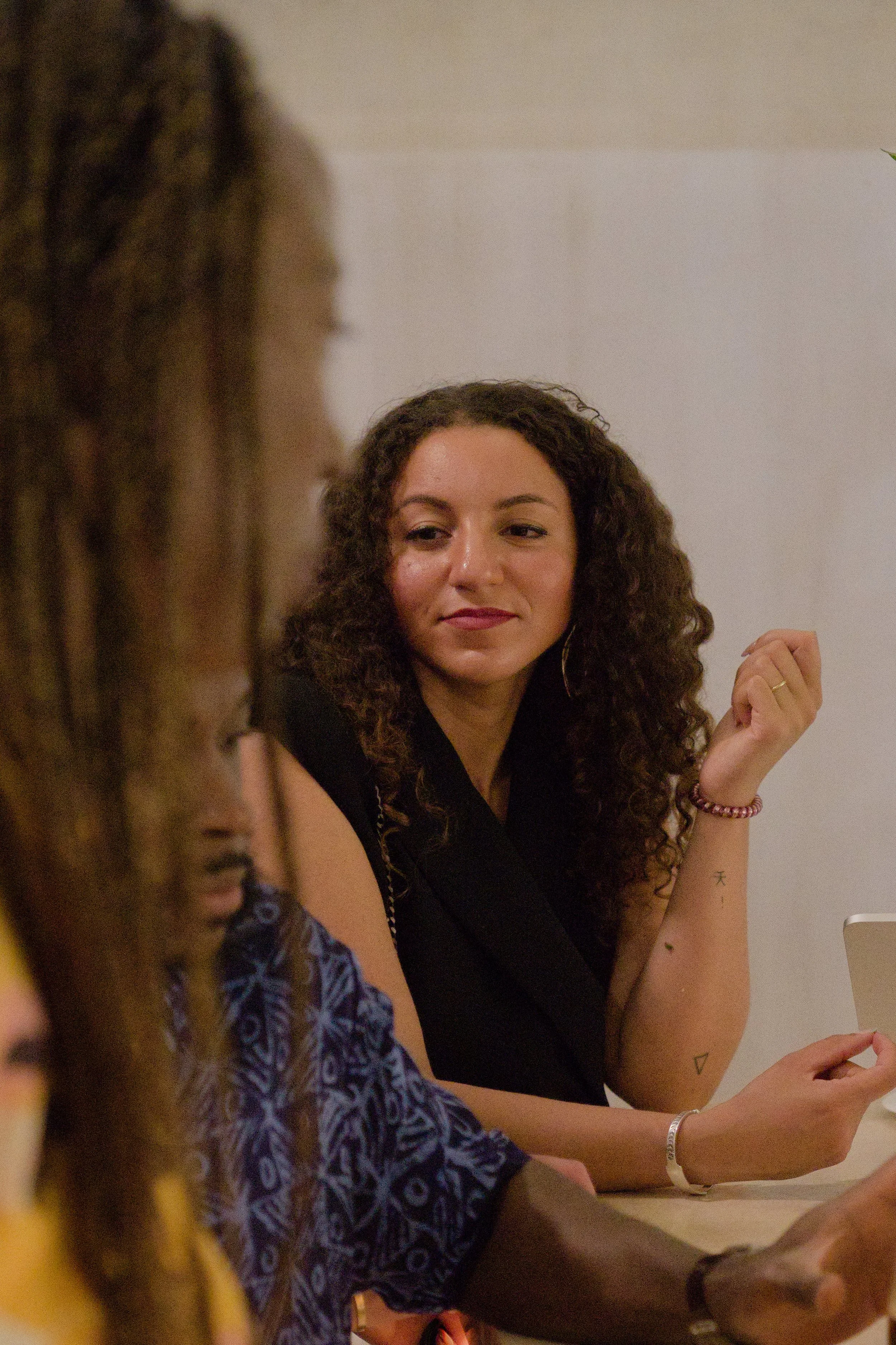 A woman with curly brown hair, wearing a black top and jewelry, looks towards a person with braided hair and a patterned blue and black garment, sitting at a table.