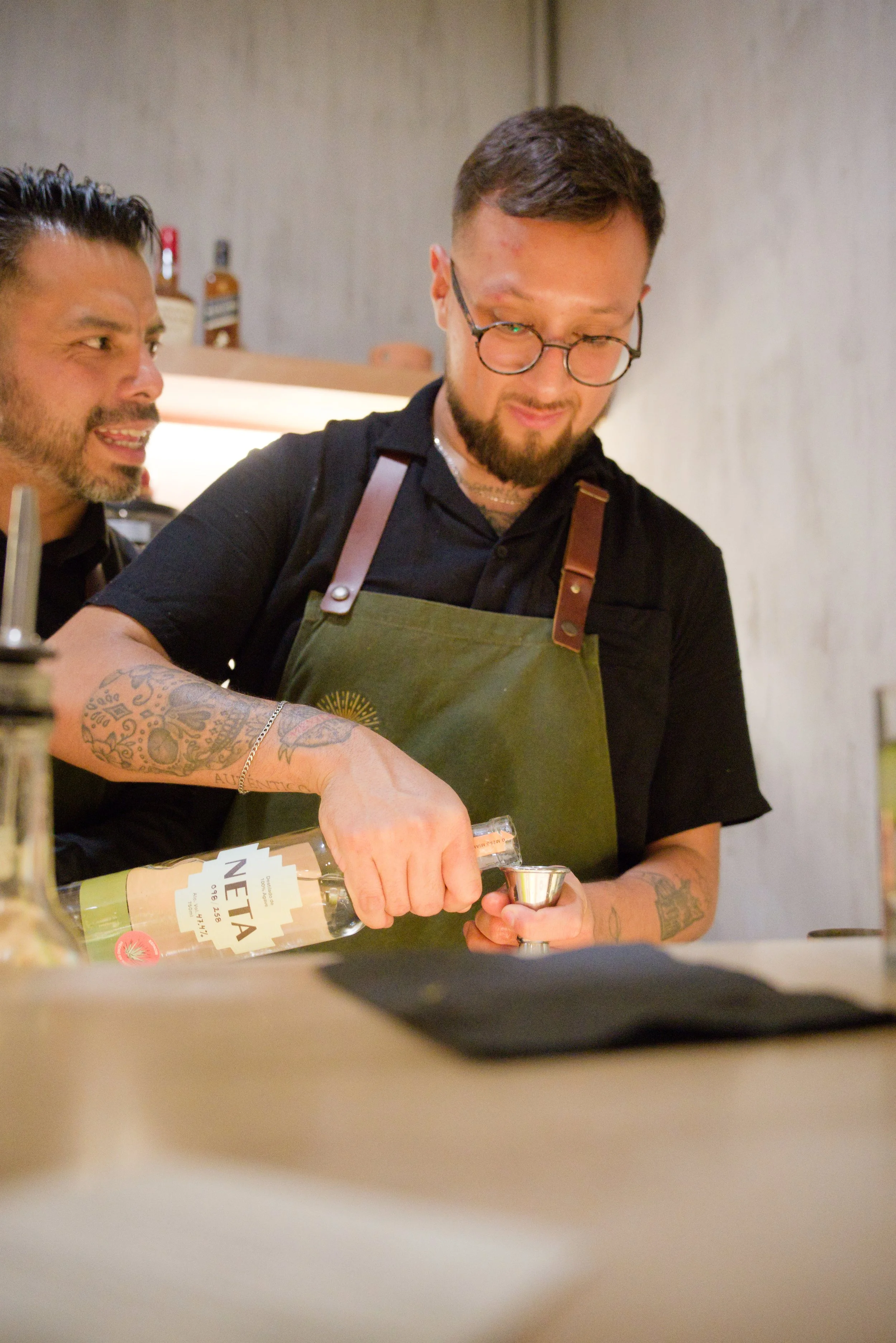 Two men preparing drinks in a bar or kitchen, one pouring a clear alcohol into a shot glass, both smiling, with bottles in the background.
