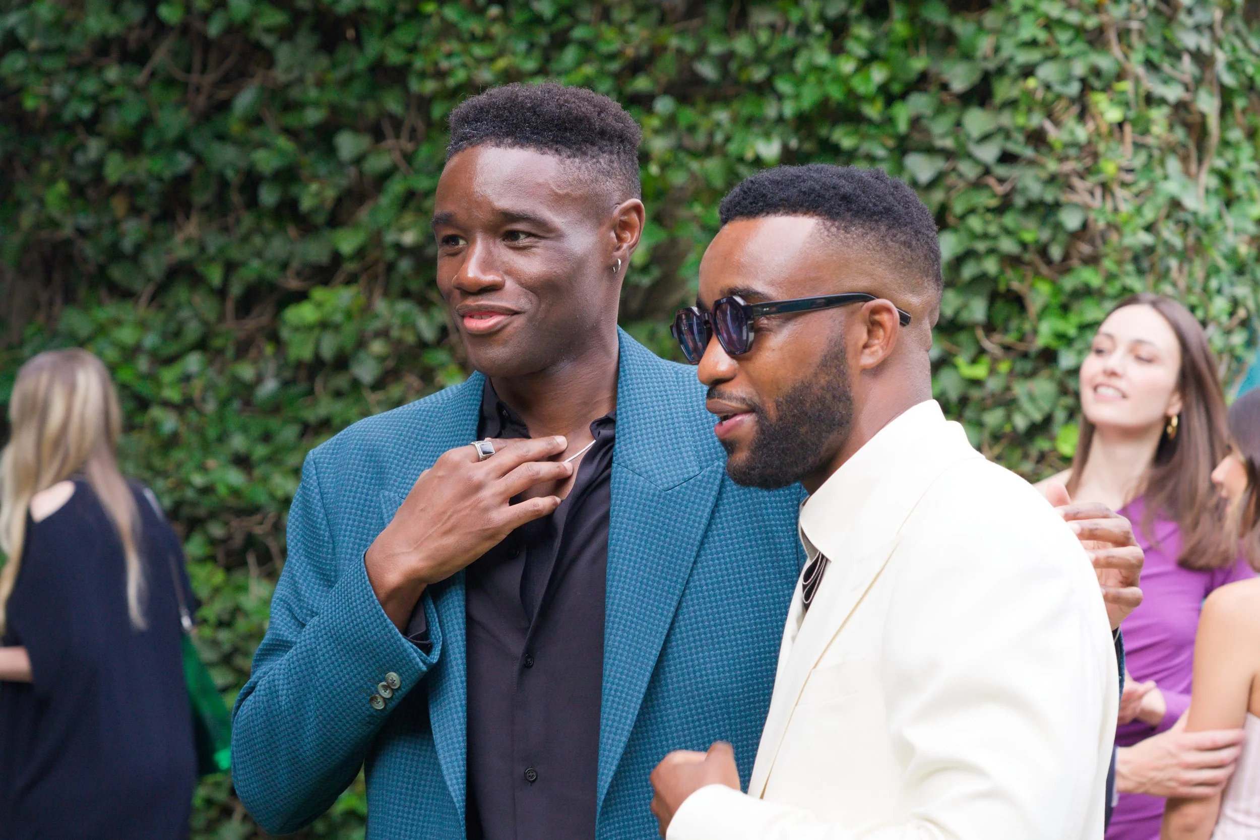 Two men socializing at an outdoor event, one in a blue blazer and the other in a white suit with sunglasses, with a green leafy background and women in dresses in the distance.