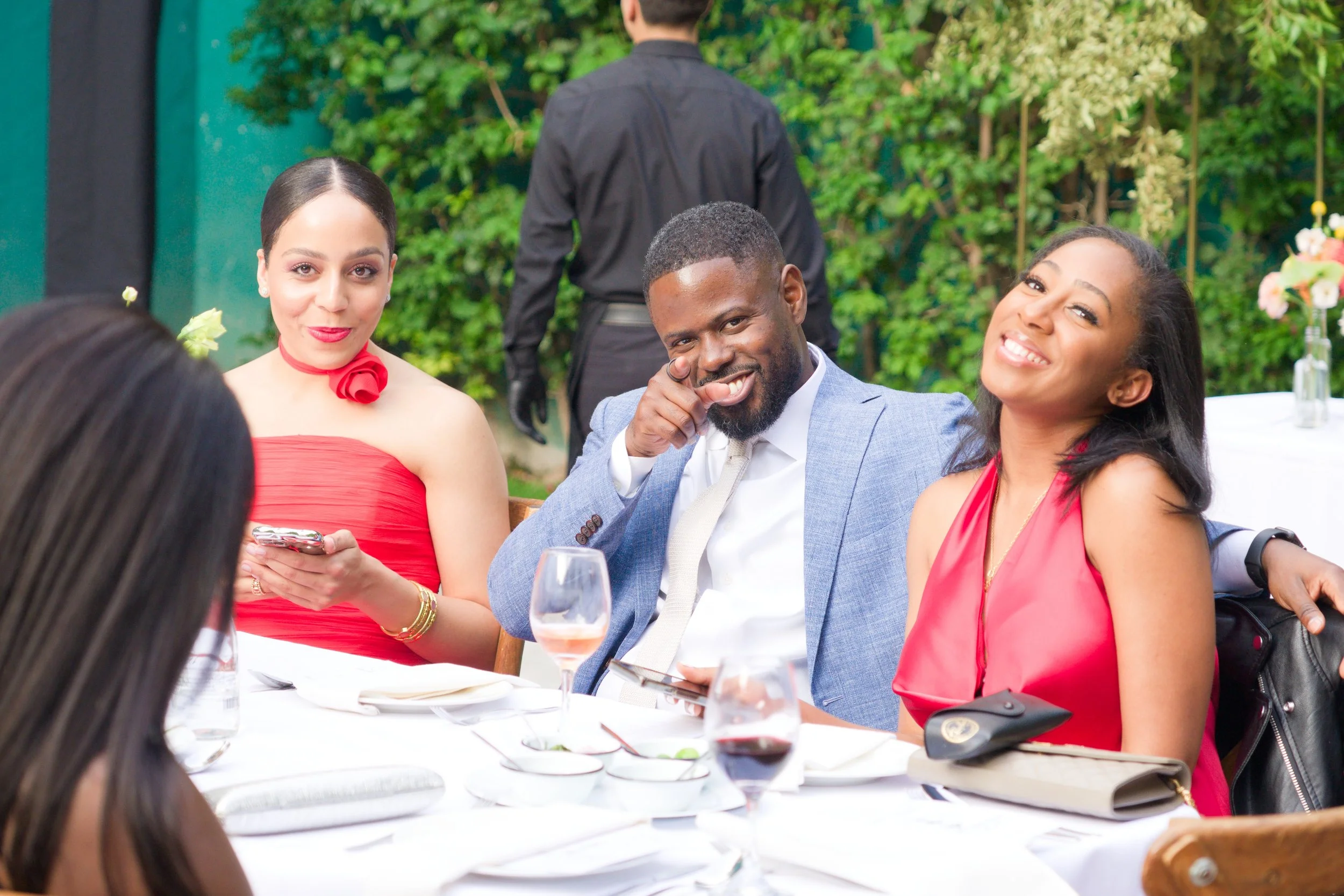 Four friends sit at a table outdoors during a celebration. A woman in a red dress and a woman in a red top are smiling, while a man in a gray suit is pointing and smiling. A woman with dark hair, seen from behind, is also present. The table has wine 