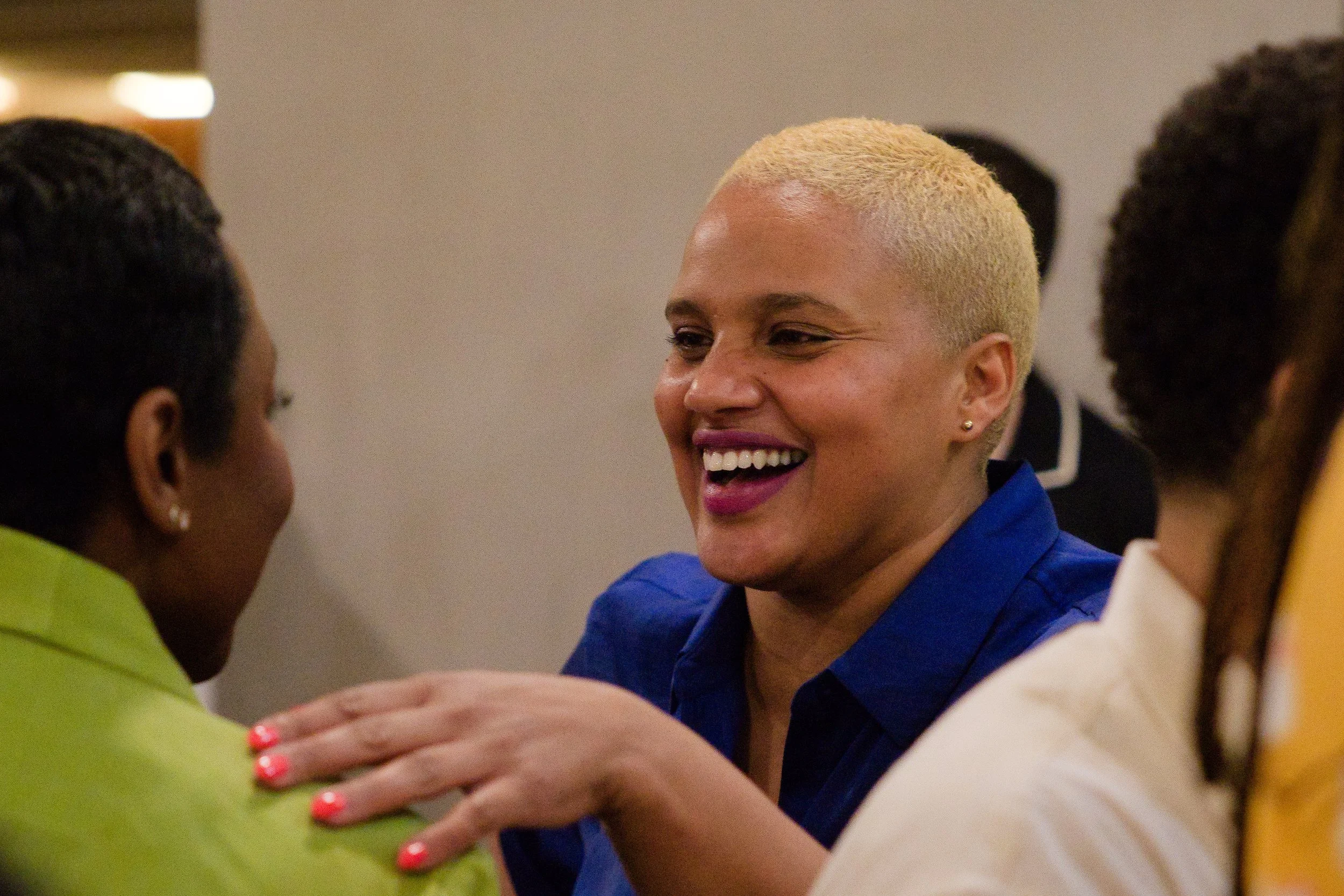 A woman with blonde hair smiling and speaking with two women, one in a green shirt and one in cream, during a social event or gathering.