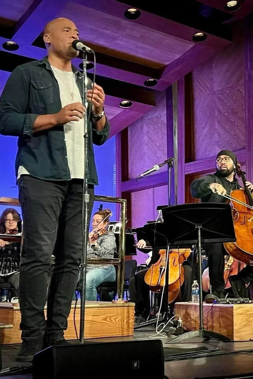 A man singing into a microphone on stage with musicians playing string instruments in the background.