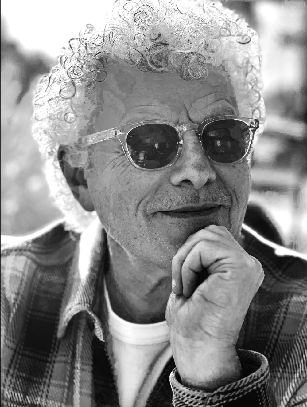 A black and white close-up of an elderly man with curly white hair, wearing sunglasses, resting his chin on his hand, smiling.