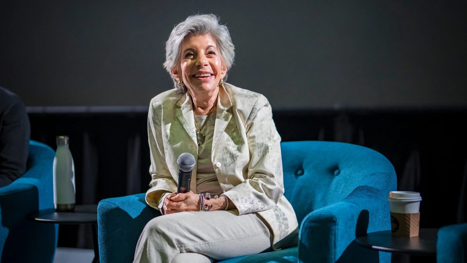 An elderly woman with gray hair smiling and holding a microphone, sitting on a blue armchair during an event.