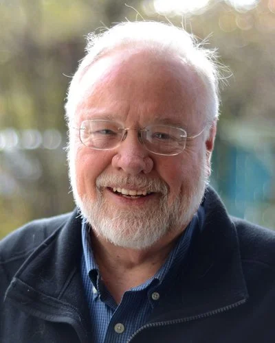A smiling older man with white hair and a beard, wearing glasses, a blue shirt, and a black jacket outdoor with blurred trees in the background.