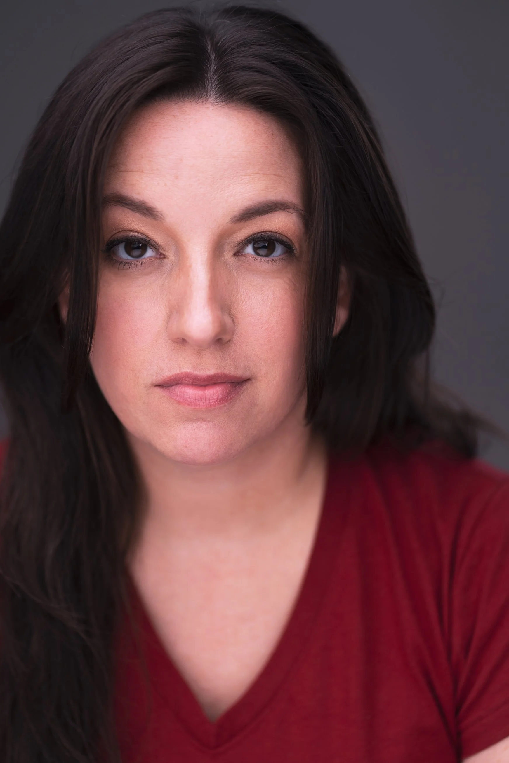 Close-up portrait of a woman with dark brown hair, light makeup, and a neutral expression, wearing a red shirt, against a dark gray background.
