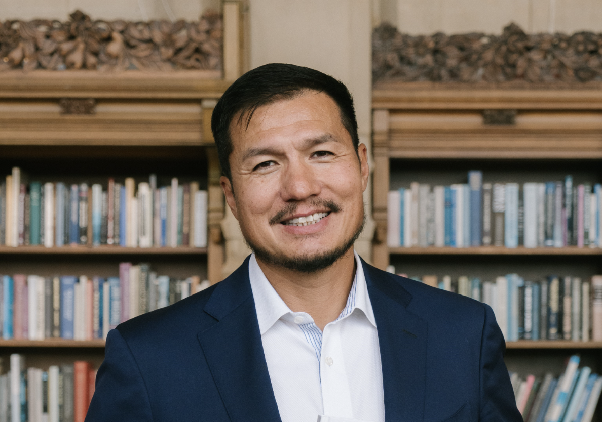 A man with short dark hair, a beard, and a mustache is smiling at the camera. He is wearing a dark blue suit jacket and a white dress shirt. In the background, there are wooden bookshelves filled with various books.