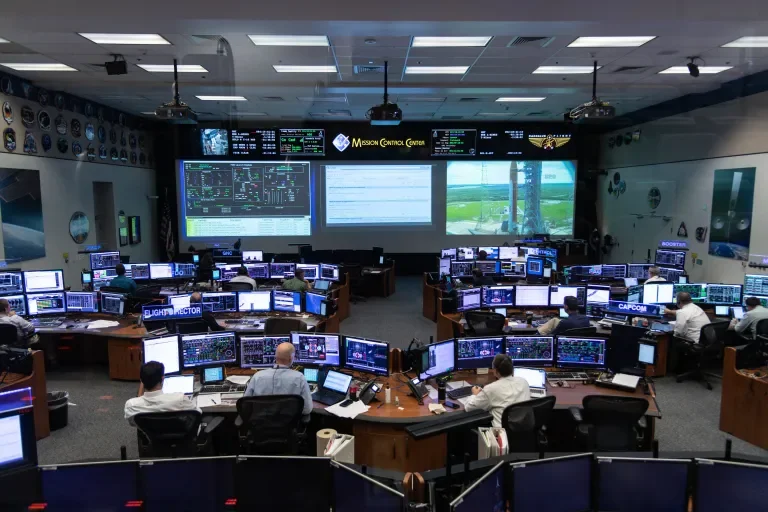 Mission Control Center with multiple employees working at computer stations, large screens displaying data, maps, and satellite images on the wall.