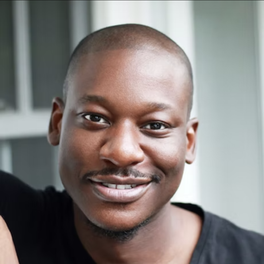 Close-up of a smiling Black man with a shaved head, wearing a black shirt, standing in front of a window with a white frame.