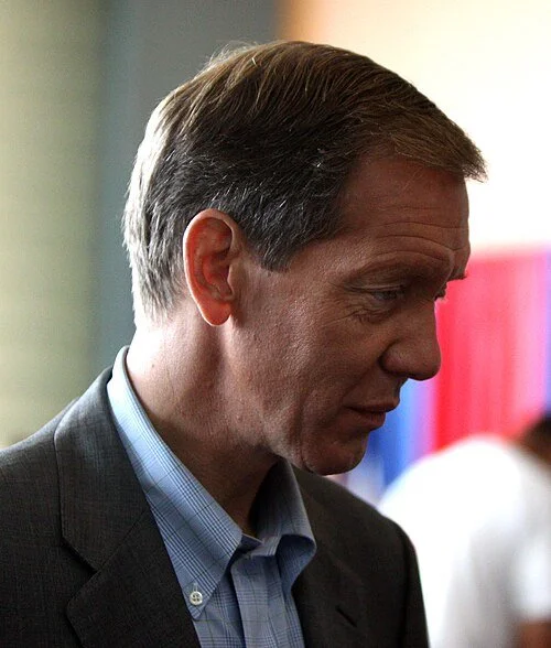A middle-aged man with short brown hair, wearing a suit and blue checkered shirt, looking down with a somber expression in an indoor setting.