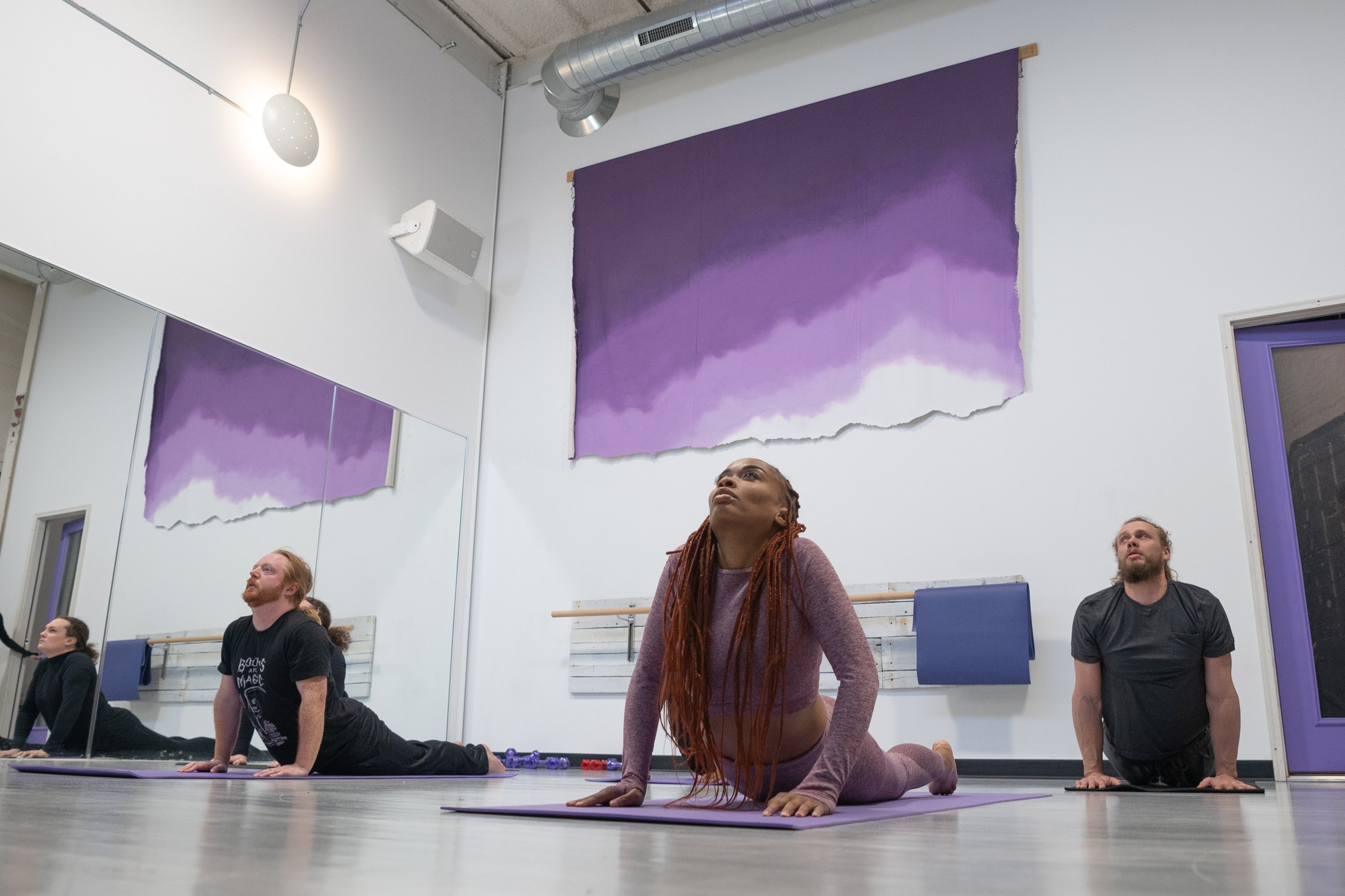 Group of people practicing yoga in a studio, sitting on purple and black yoga mats, doing a yoga pose