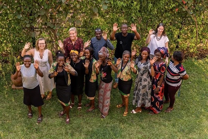 Group of smiling women and men from the brand Ndara waving, standing outdoors on grass with lush green foliage in the background.