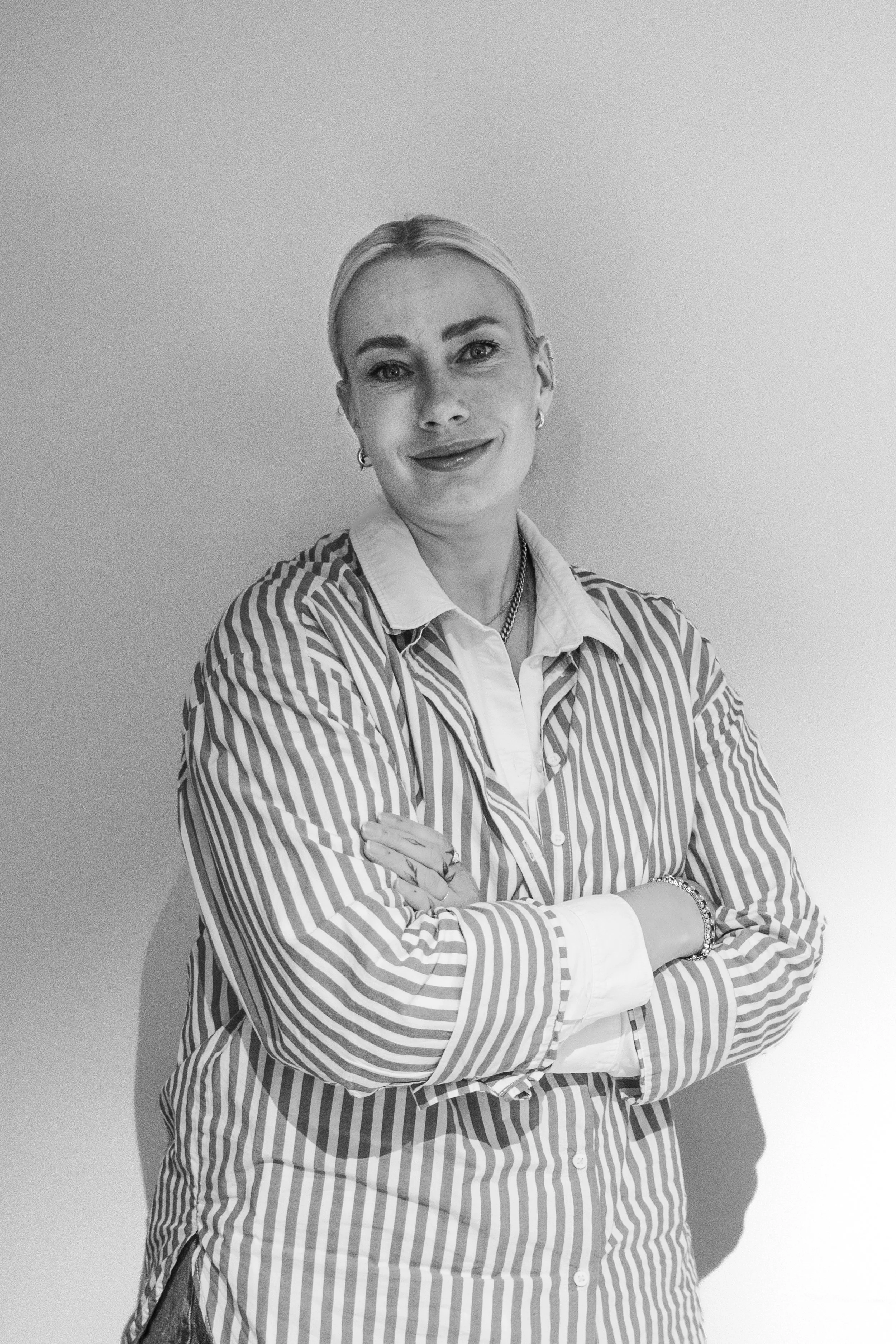A woman standing against a plain wall, with her arms crossed and smiling at the camera. She has blonde hair and is wearing a striped long-sleeve shirt over a collared blouse, accessorized with earrings, a necklace, and a bracelet.