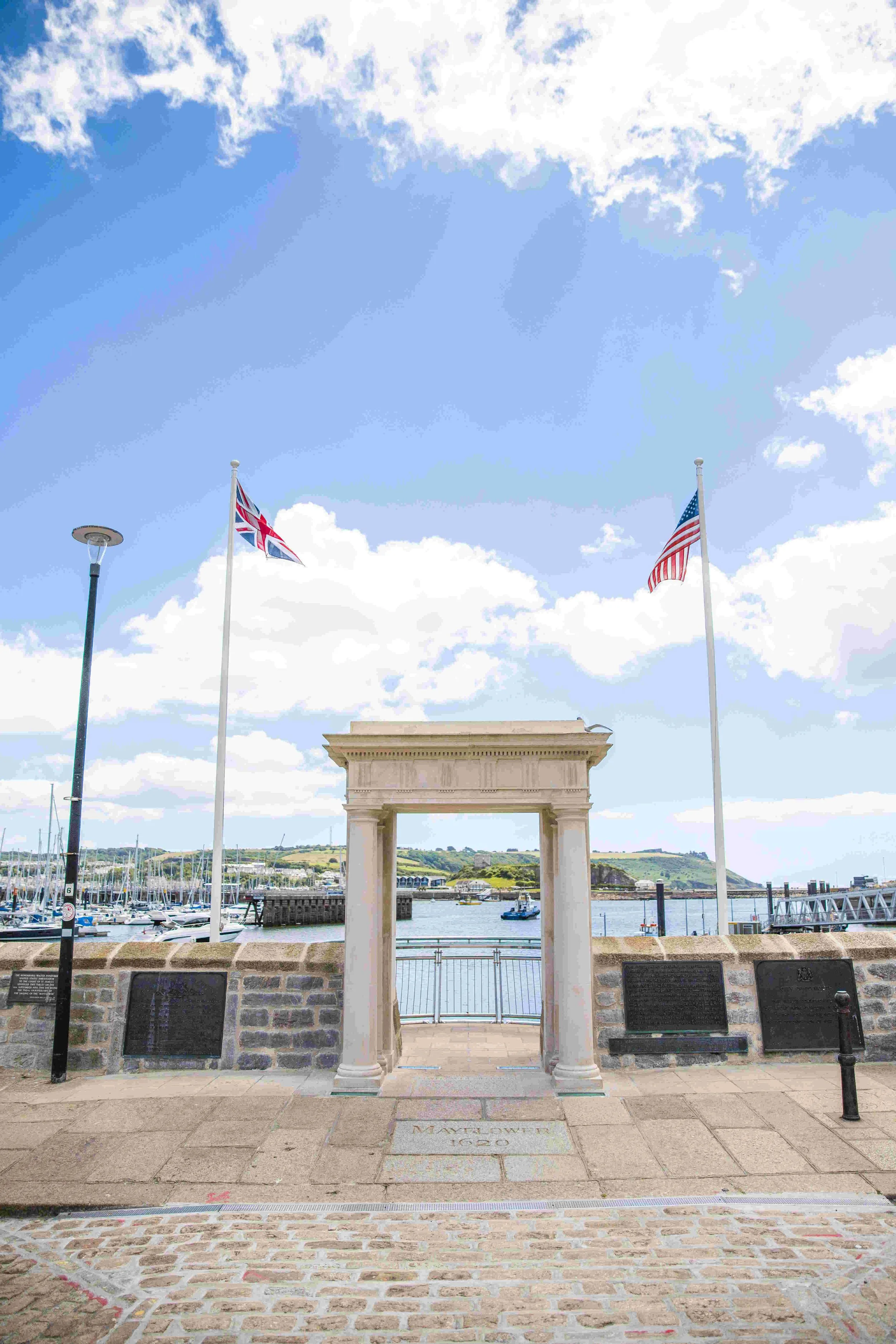 A waterfront scene with two flagpoles, one with the United Kingdom flag and the other with the United States flag, a stone archway labeled 'Mayflower 1620,' and a marina with boats, under a partly cloudy sky.