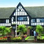 A black and white Tudor-style house with a steep roof, surrounded by green lawn and palm trees.
