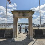 Stone archway on a waterfront with cloudy sky, people in the distance, and boats on the water