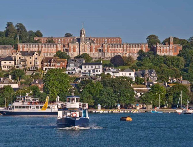 Boat sailing on blue water with colorful buildings and a large historic brick building atop a hill in the background.