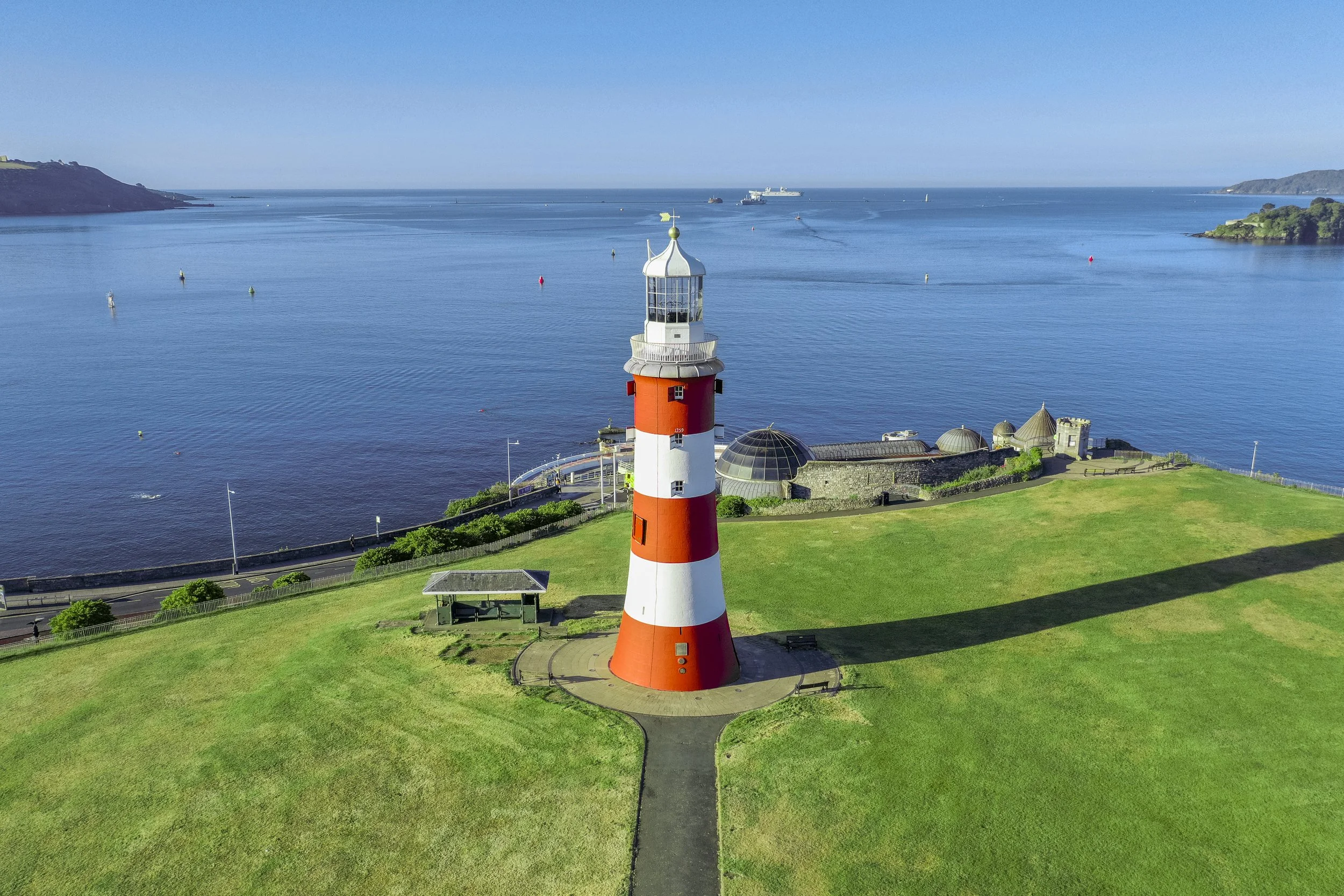 A red and white lighthouse on a grassy area by the water, with a stone building with domed and pointed roofs nearby, and ships visible in the blue ocean in the background.