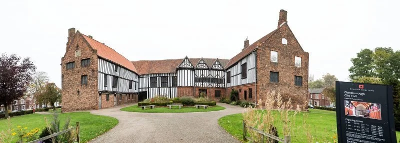 Historical building with black and white half-timbered facade, brick walls, red tile roof, surrounded by a grassy lawn and pathways, with a modern informational sign nearby.