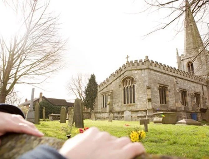 A historic stone church with a steeple and a graveyard in the foreground, with a person's arm visible in the bottom of the image.