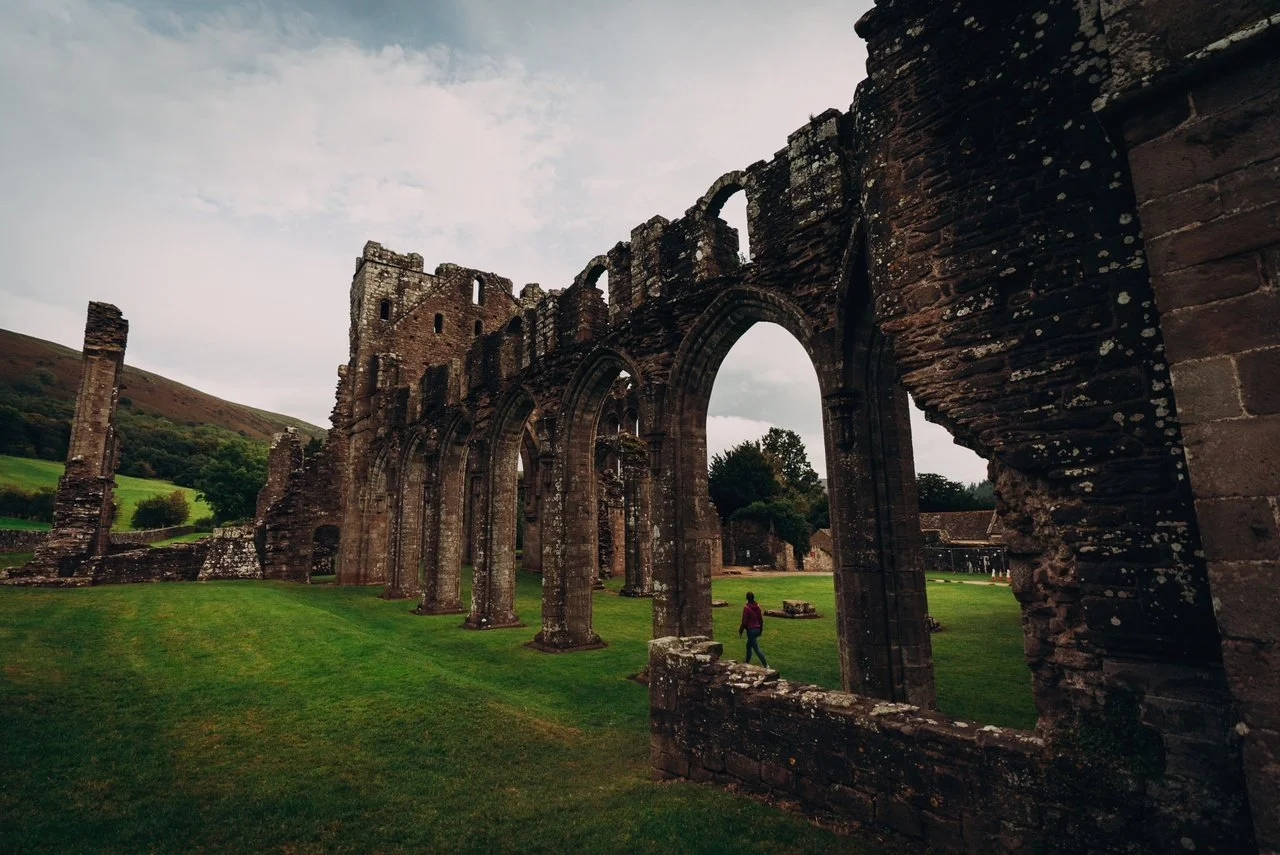 Ancient stone ruins of a historical building with arched windows and walls, set in a grassy landscape with a person walking nearby, under an overcast sky.