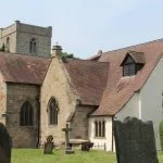 Historic church with stone walls and a red-tiled roof, surrounded by a grassy area and trees.