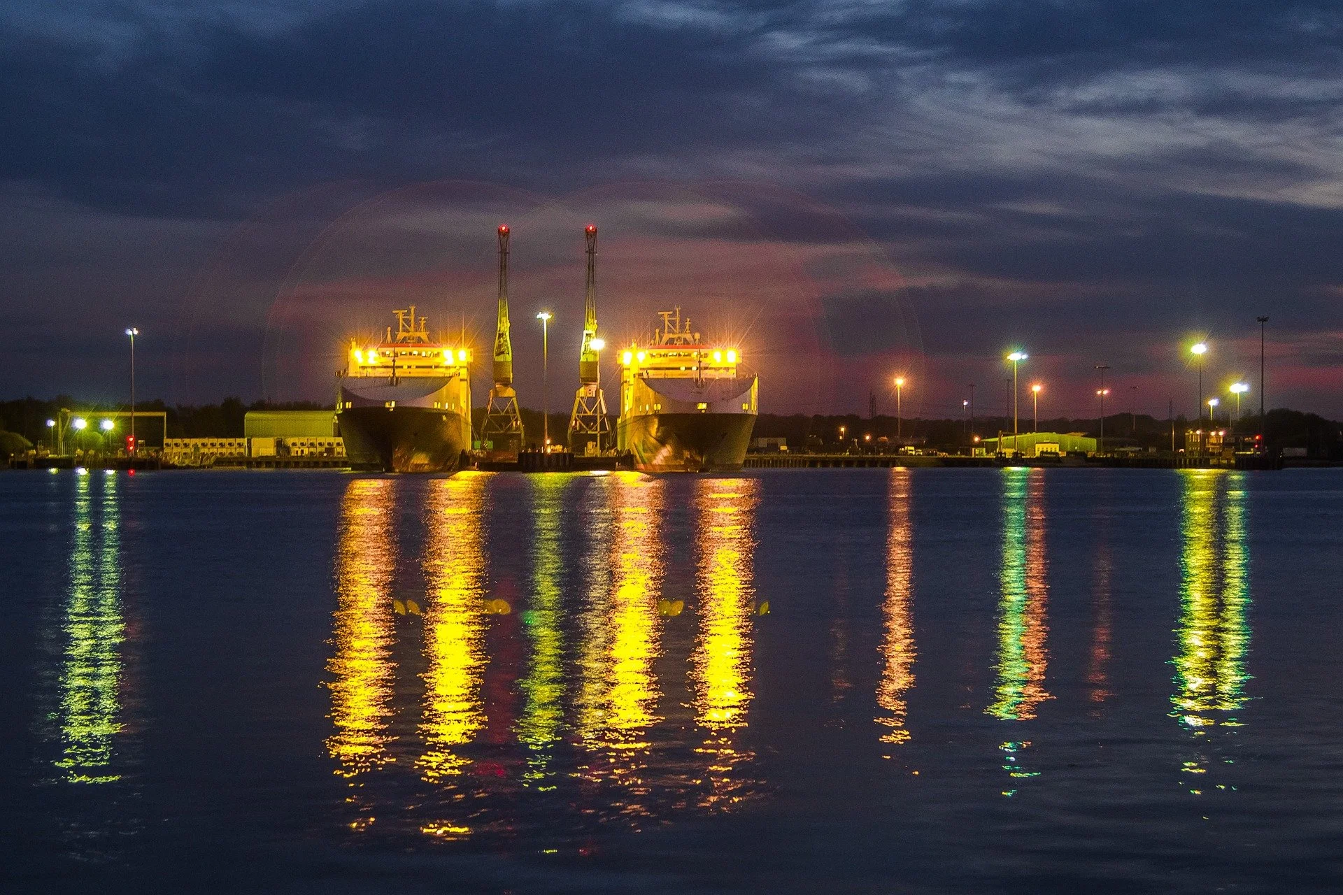 Two large ships docked at a port during twilight with their lights reflecting on the water.