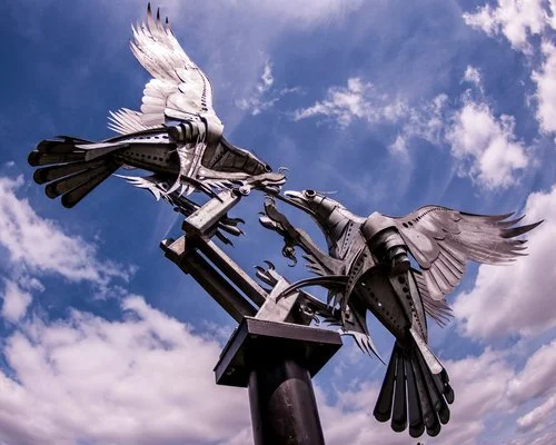 A metal sculpture of two birds made from recycled materials, with wings spread, set against a partly cloudy sky.