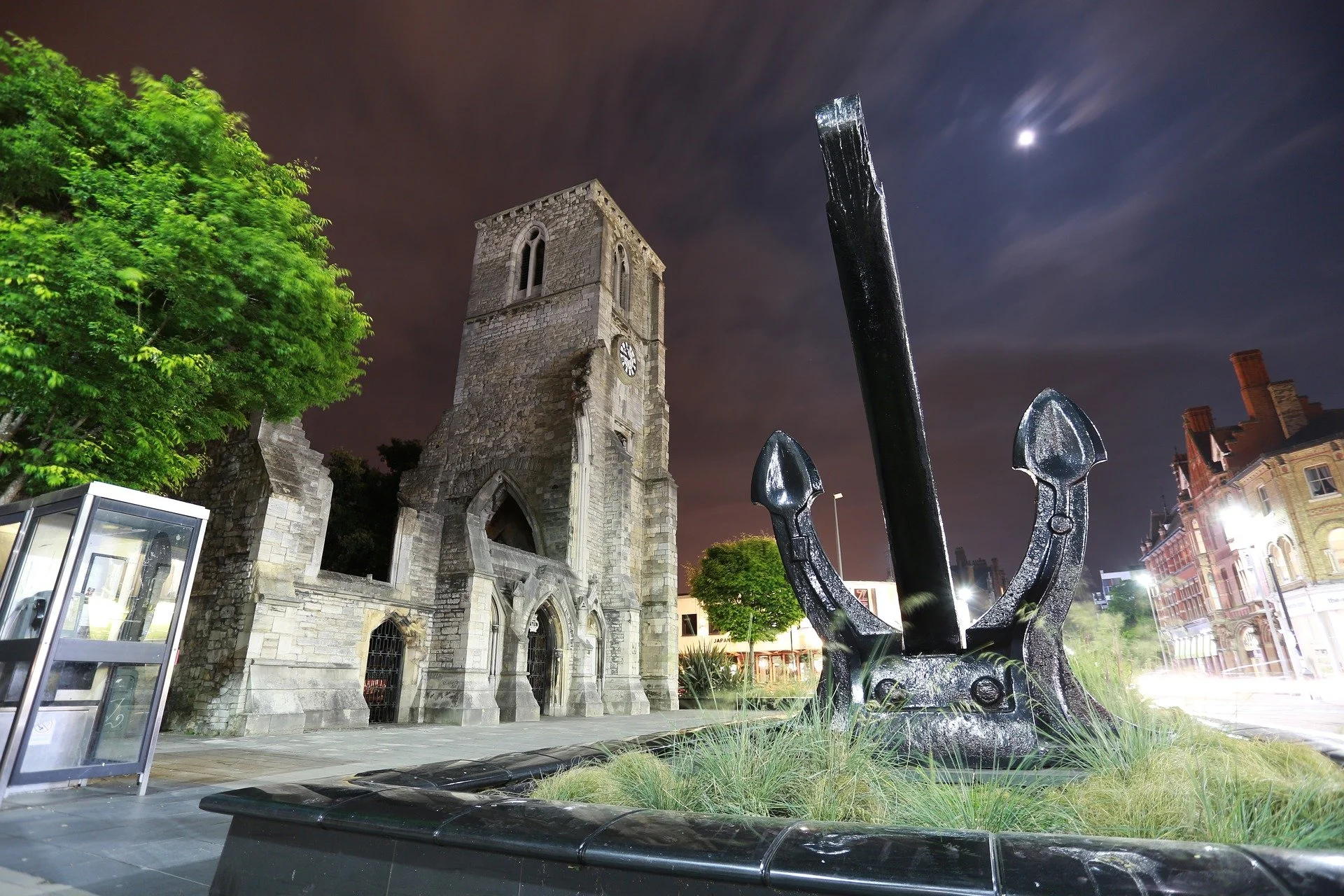 Nighttime scene of a historic stone church with a clock tower, a large black anchor sculpture in a grassy area, surrounded by trees and city buildings, with a moon in the cloudy sky.