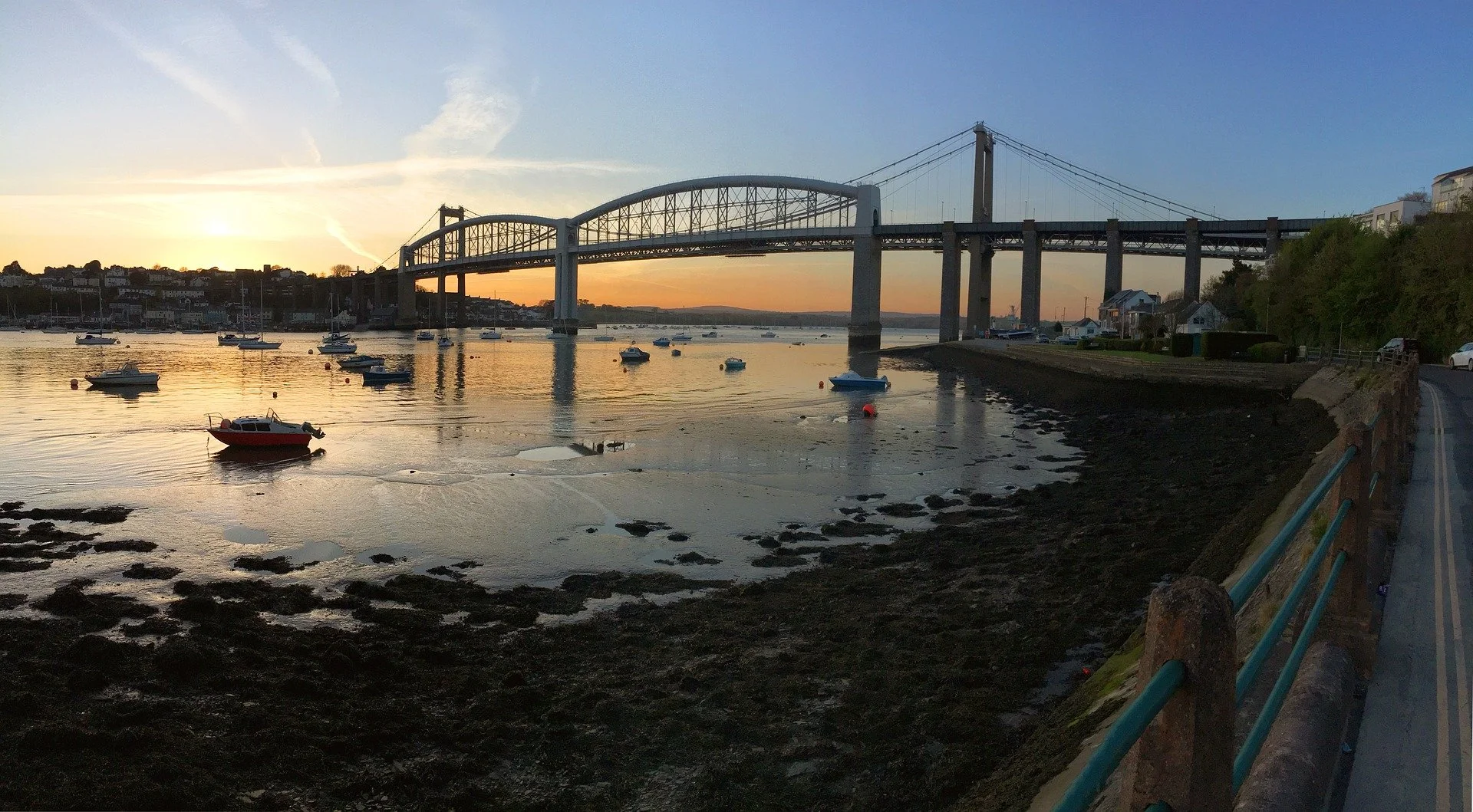 Sunset over a calm bay with boats anchored, a bridge spanning across the water, and houses along the shoreline on a clear evening.