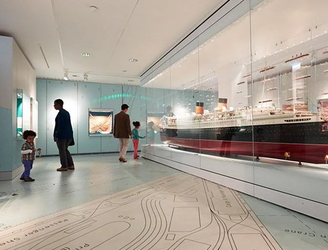 A museum exhibit featuring a large model ship enclosed in glass, with visitors observing it.