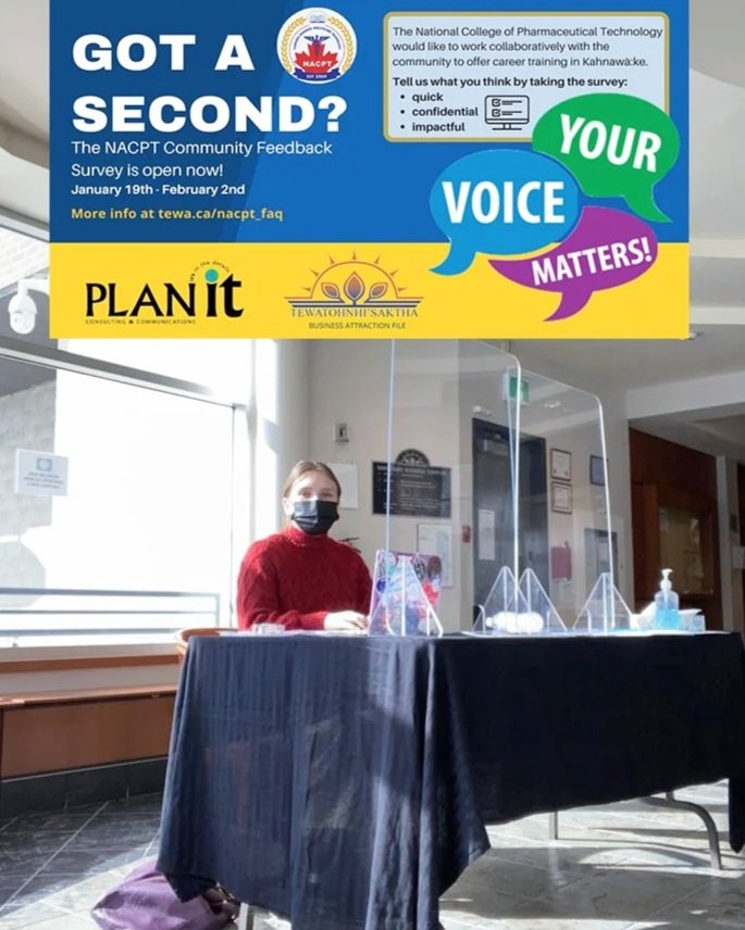 A woman wearing a red sweater and a black face mask sitting behind a table with a black cloth cover and clear plastic dividers in a room with a window and a wall with signs. Above her, a blue and yellow sign promotes a survey for the NACPT community feedback, encouraging participation to amplify your voice and matter.
