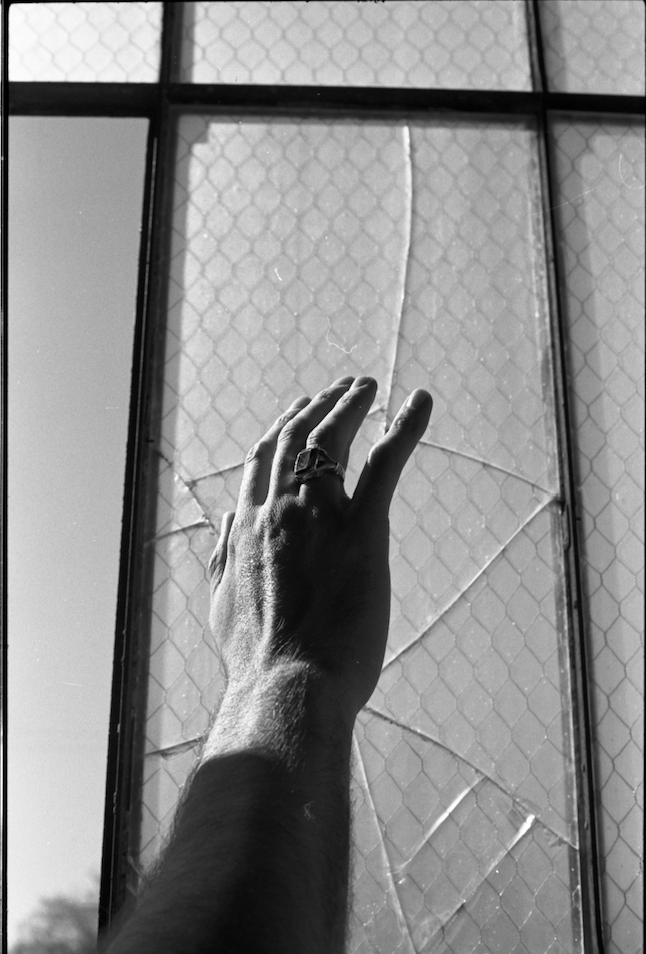Photograph of a man’s hand wearing a ring, reaching toward broken glass, illuminated by ethereal, otherworldly light.