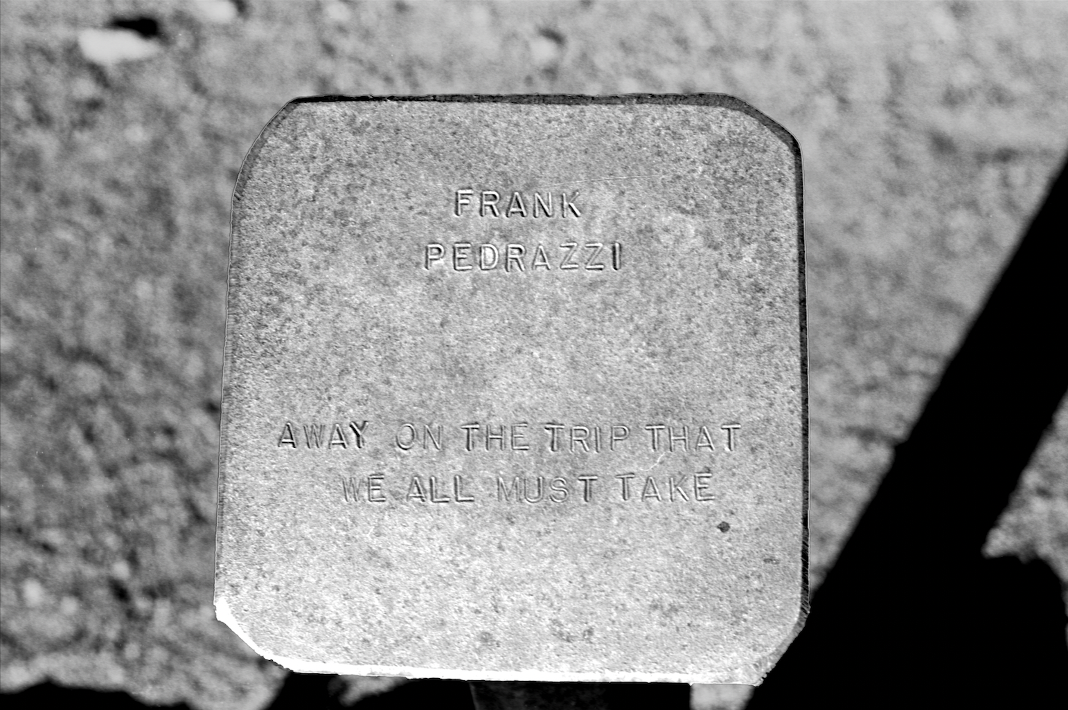 Black-and-white photograph of a metal-punched headstone in Tonopah, Nevada, behind the Clown Motel, engraved with “Away on the trip that we all must take”, its weathered form stark against the desert landscape.