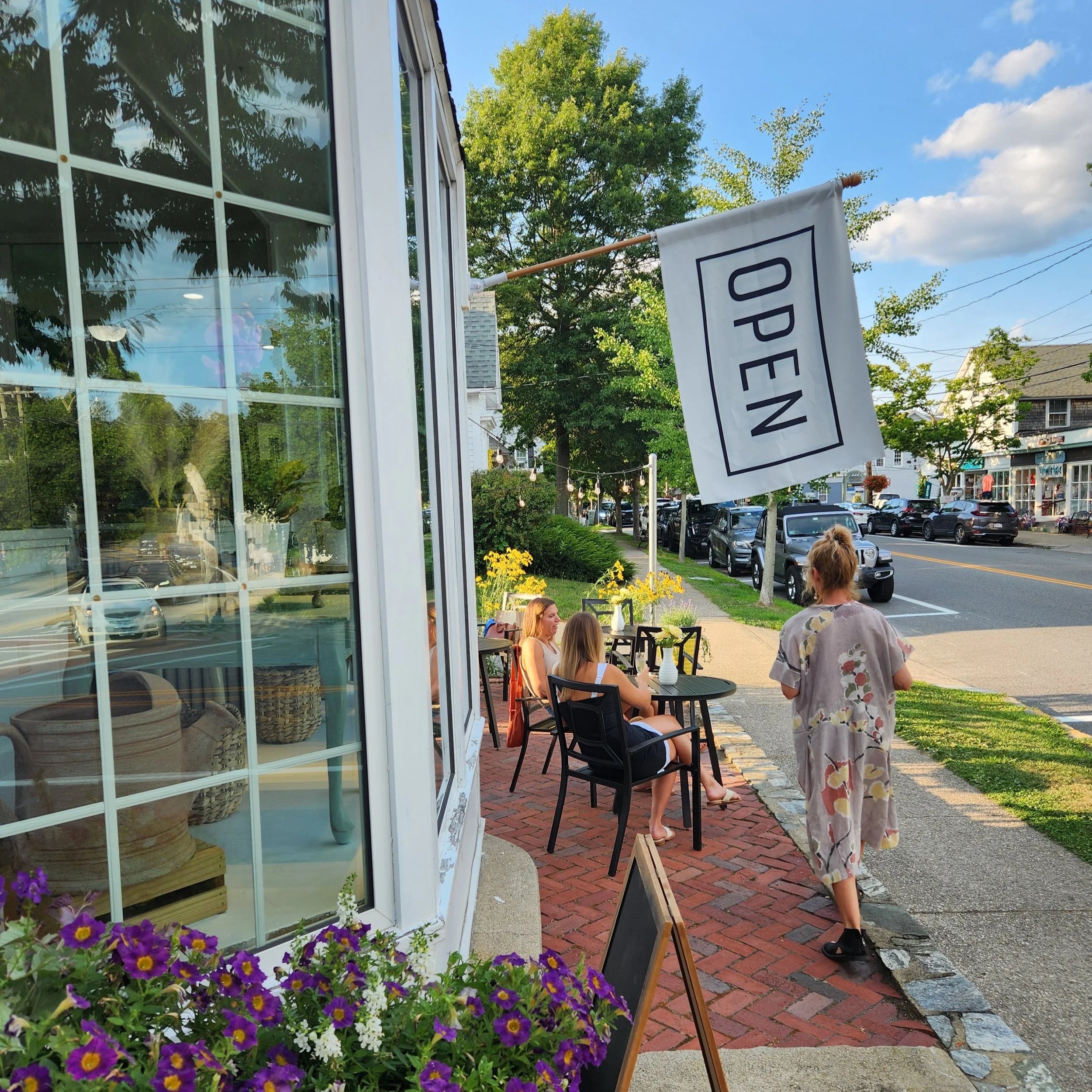 Sidewalk outdoor cafe with four women sitting and a woman walking, storefront with large glass window, flowers outside, sign hanging with 'OPEN', trees, parked cars, and houses in a small town atmosphere.