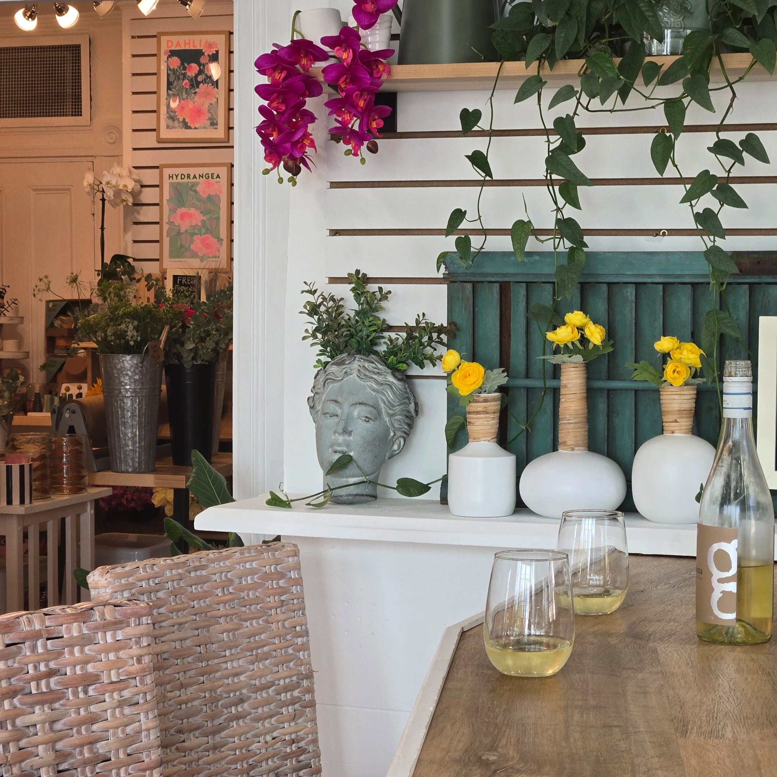Interior of a cozy cafe with decorative plants, colorful flowers, and modern art on the walls. There are two glasses of white wine and a bottle on the wooden table, with woven chairs.