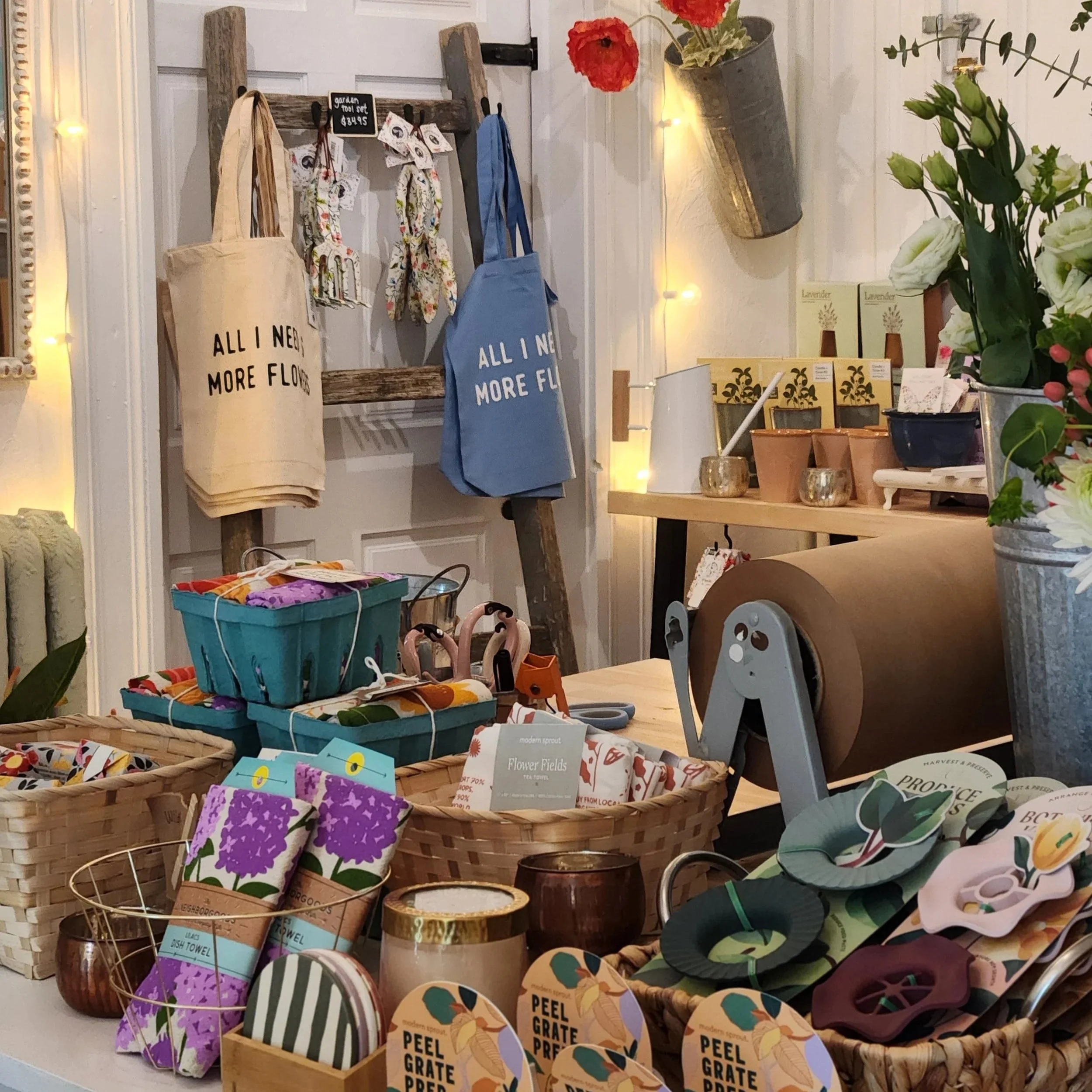 Display of various home decor and gift items in a cozy store, including tote bags, packaged soaps, candles, rolled paper, and small decorative objects, with warm lighting and floral accents.
