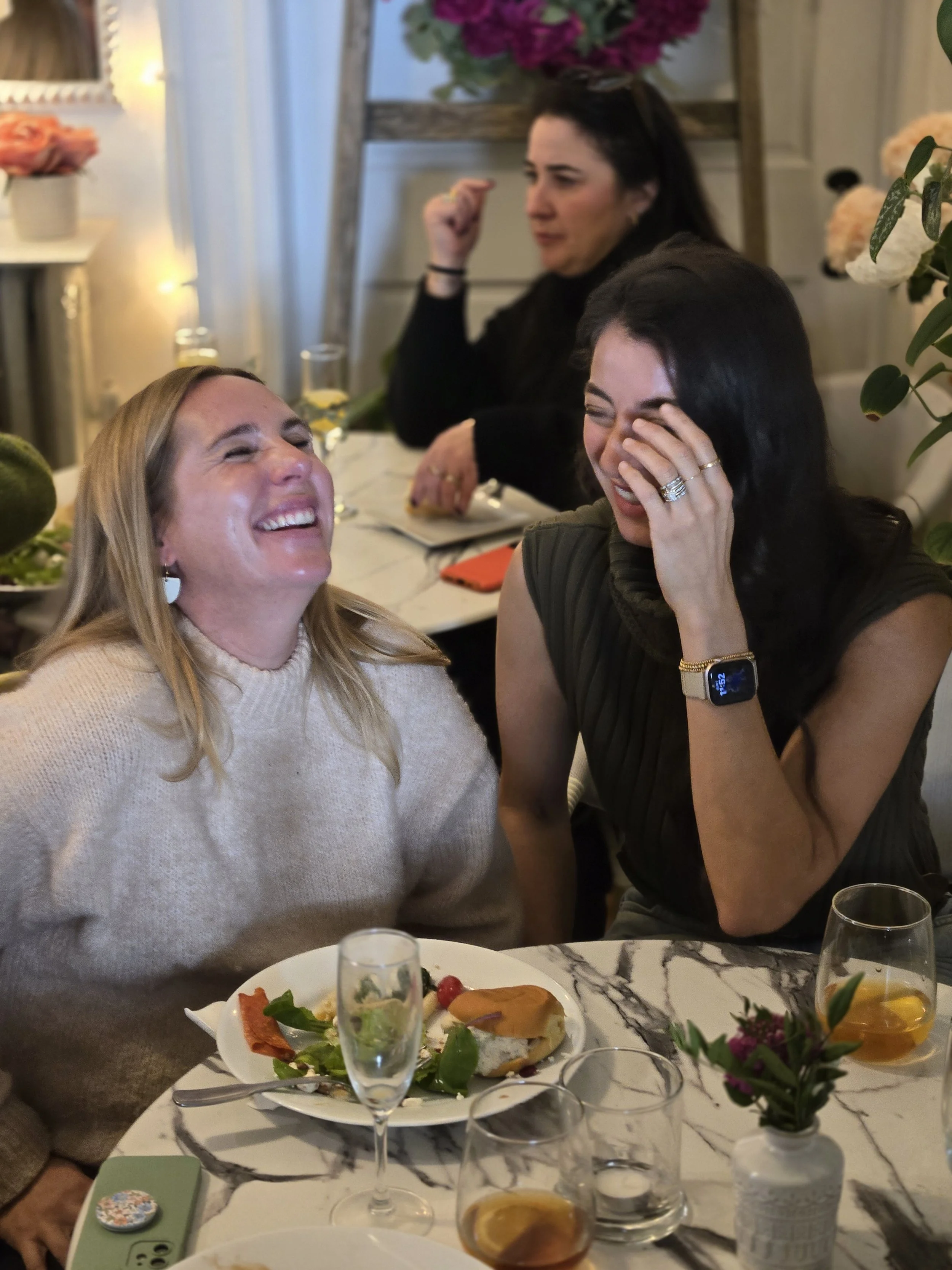 Two women laughing and smiling at a dinner table, with one covering her face with her hand. There are plates of food, glasses of drinks, and a small flower arrangement on the table. In the background, another woman is sitting with her hand raised.