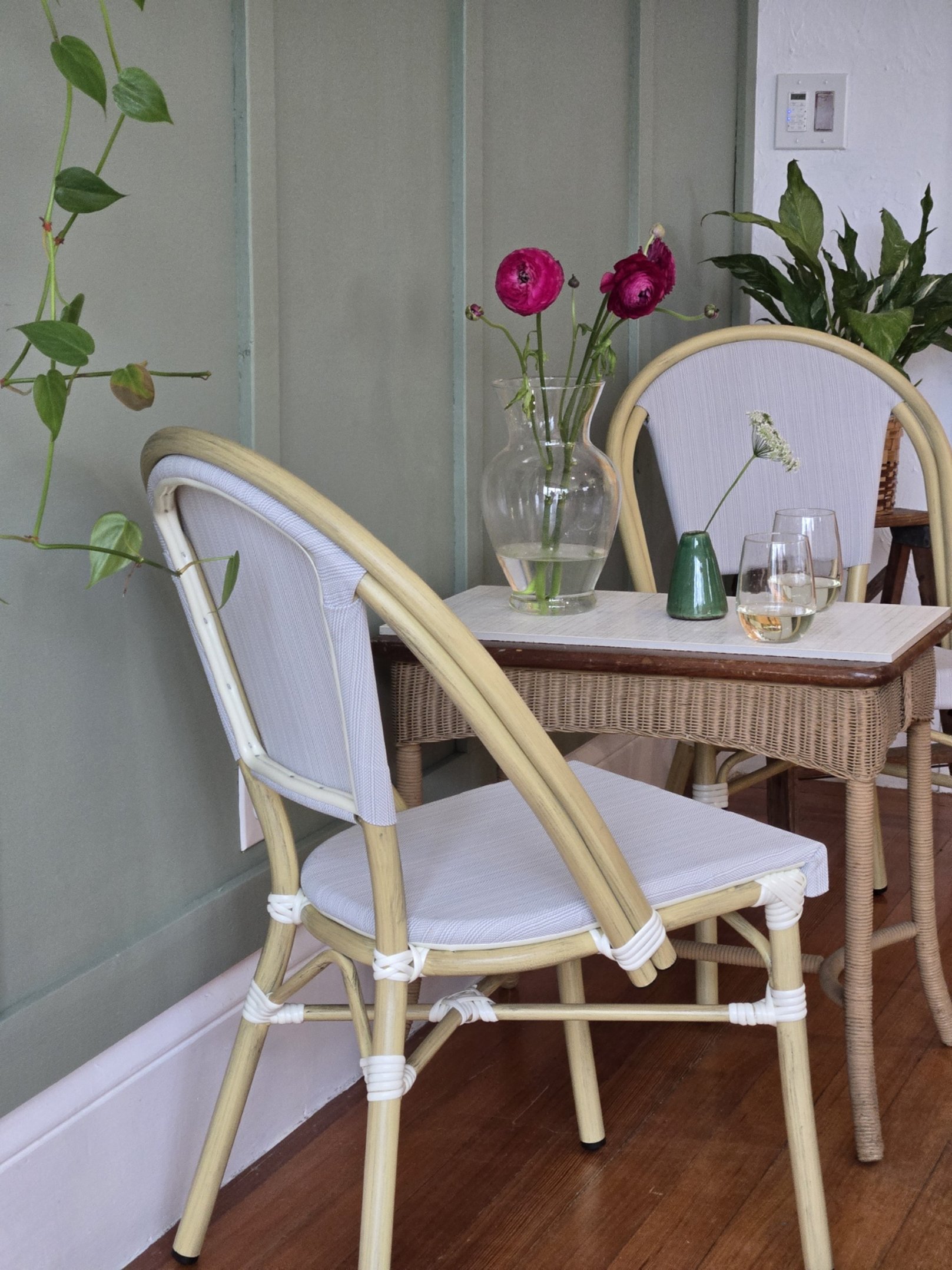 A table with a glass vase of pink flowers, two glasses of wine, and a small green vase, with two white cushioned chairs and potted plants in the background.