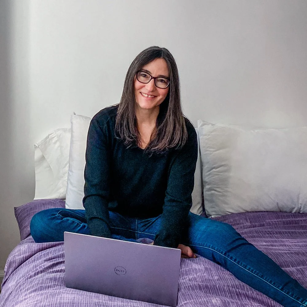 Shannah Albert Squarespace Web Designer, sitting cross-legged on a bed with a purple bedspread and white pillows, smiling at the camera with a laptop in front of her