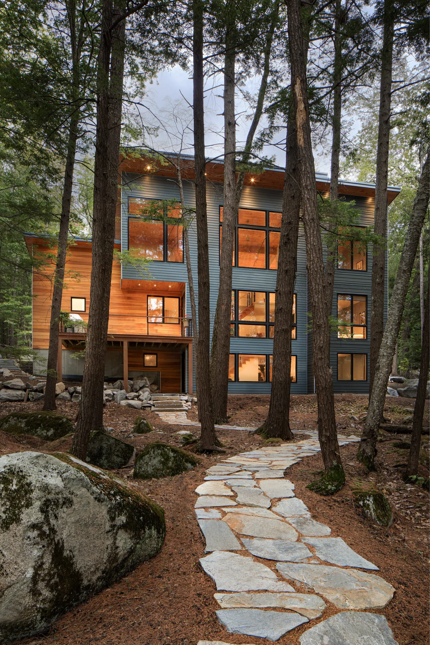 Modern multi-story house with large windows, surrounded by tall trees and a stone pathway leading up to the entrance in a forested area.