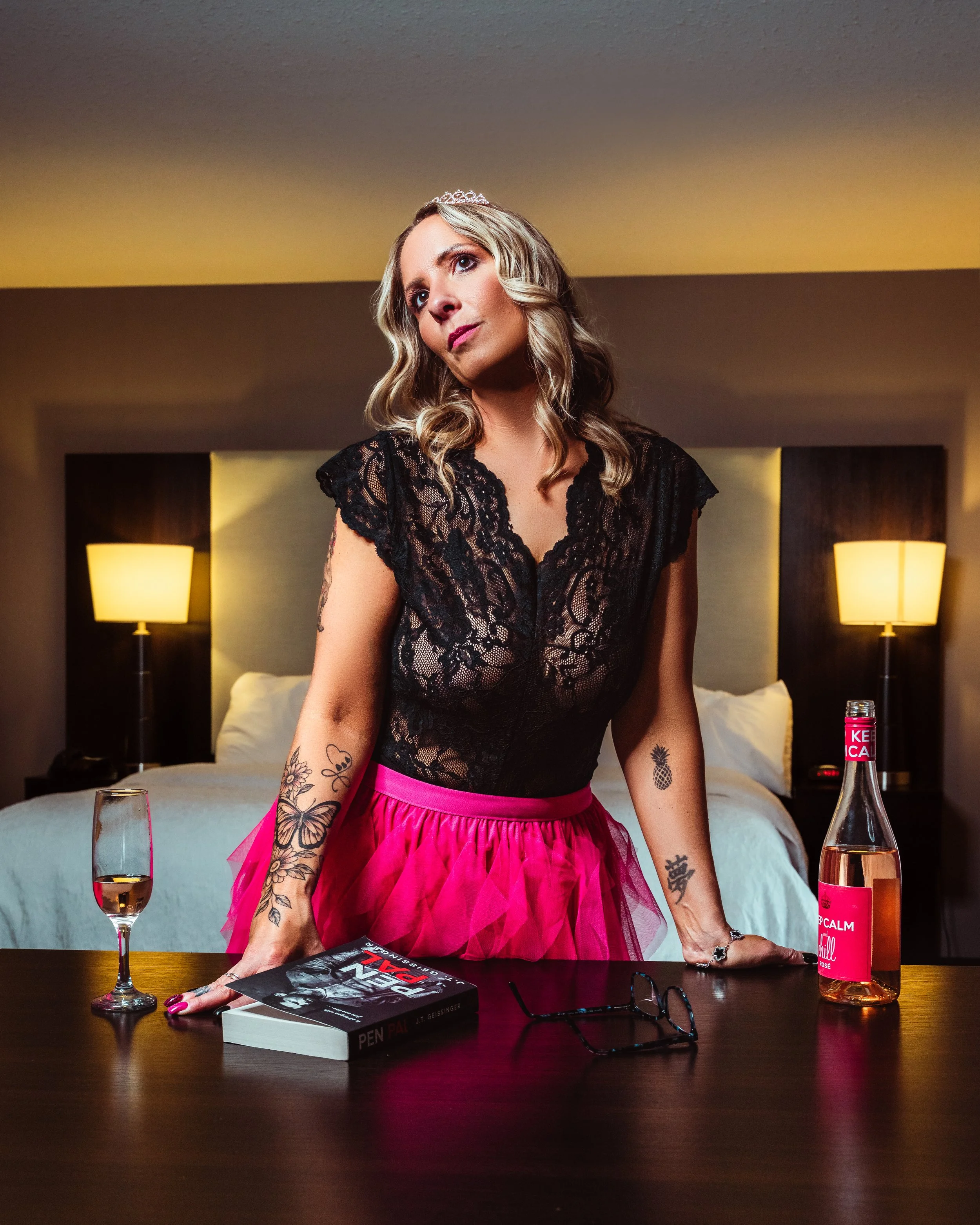 Woman with wavy blonde hair wearing a tiara, black lace top, and a pink skirt standing behind a dark wooden table in a hotel room, with two lamps on the wall behind her, a glass of wine, a book titled "Pen Pal" by J.T. Geissinger, a pair of glasses, 