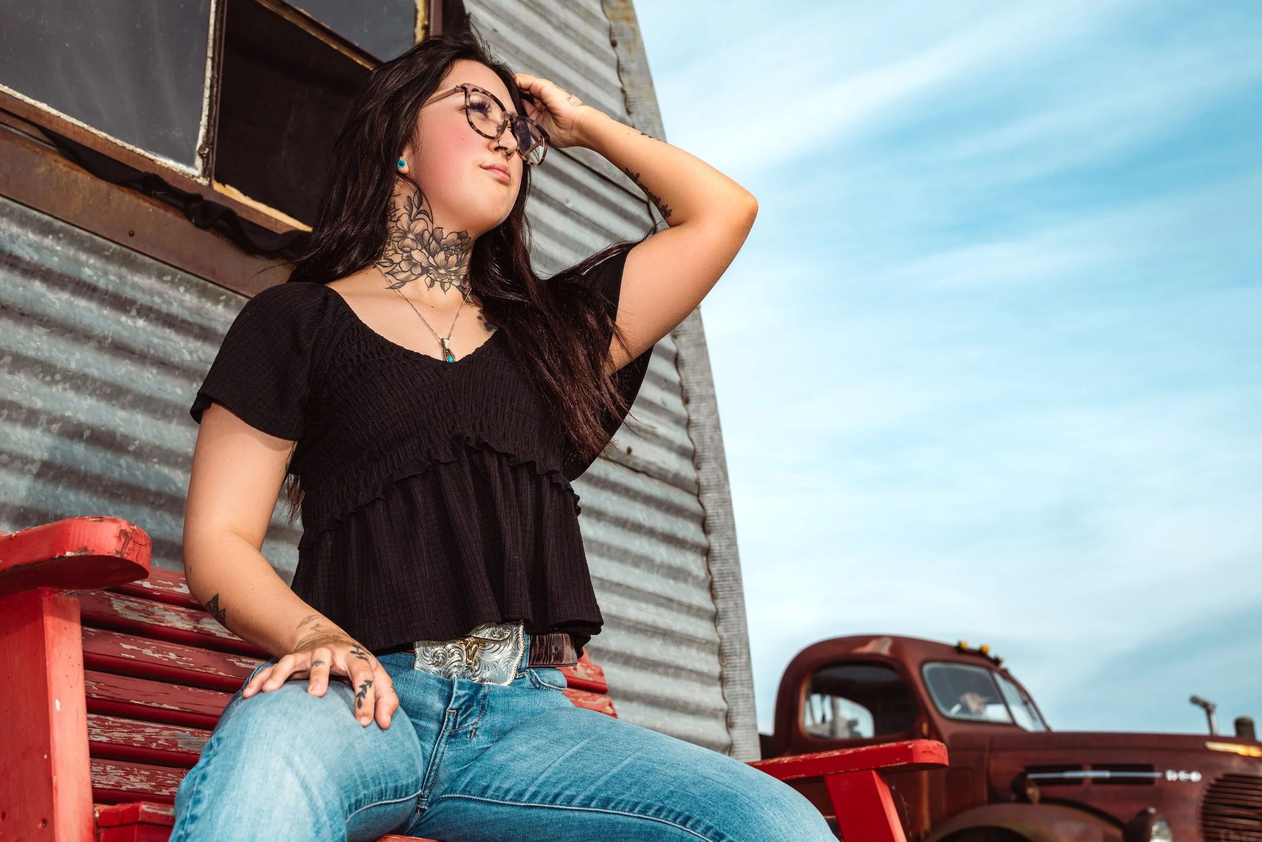 A young woman with long dark hair, tattoos, and glasses sitting on a red wooden bench against a corrugated metal wall. She is wearing a black top and light blue jeans, with her hand resting on her knee. She appears relaxed, with her eyes closed and a